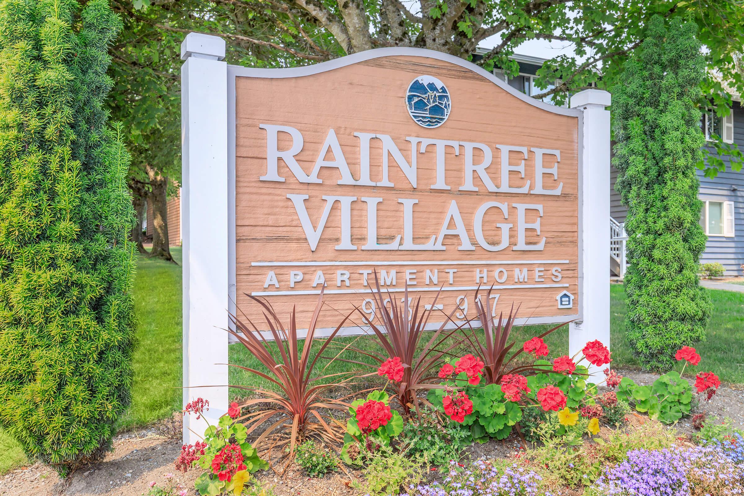 A wooden sign for Raintree Village Apartment Homes, featuring the name prominently displayed. Surrounding the sign are decorative plants and vibrant flower beds with red flowers, set against a backdrop of green grass and trees. The overall scene conveys a welcoming atmosphere.