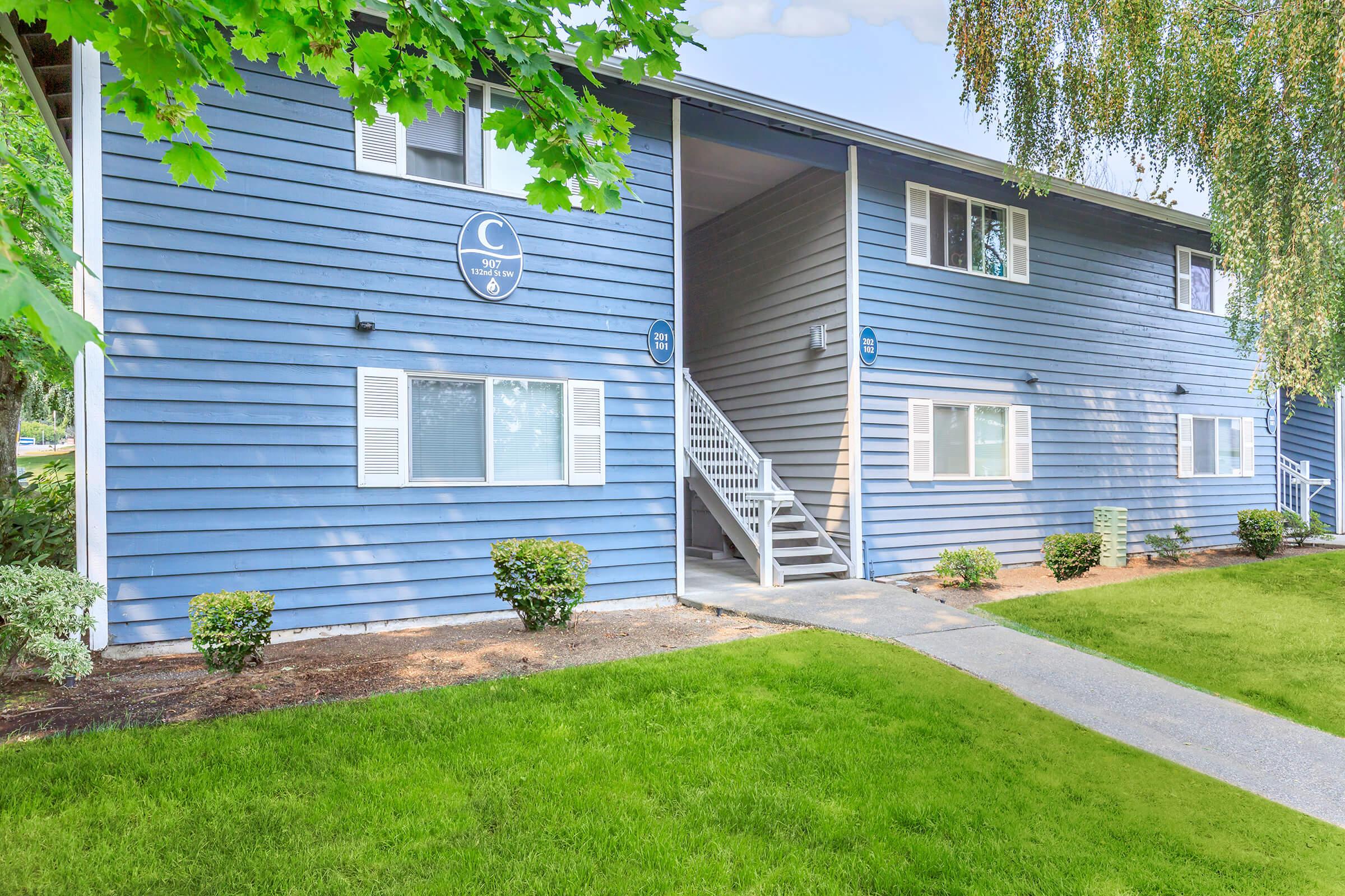 A multi-unit residential building with a blue exterior, featuring a central staircase leading to upper floors. The landscaping includes manicured grass and small shrubs, with windows on both levels. A logo or identifier is visible above the entrance.