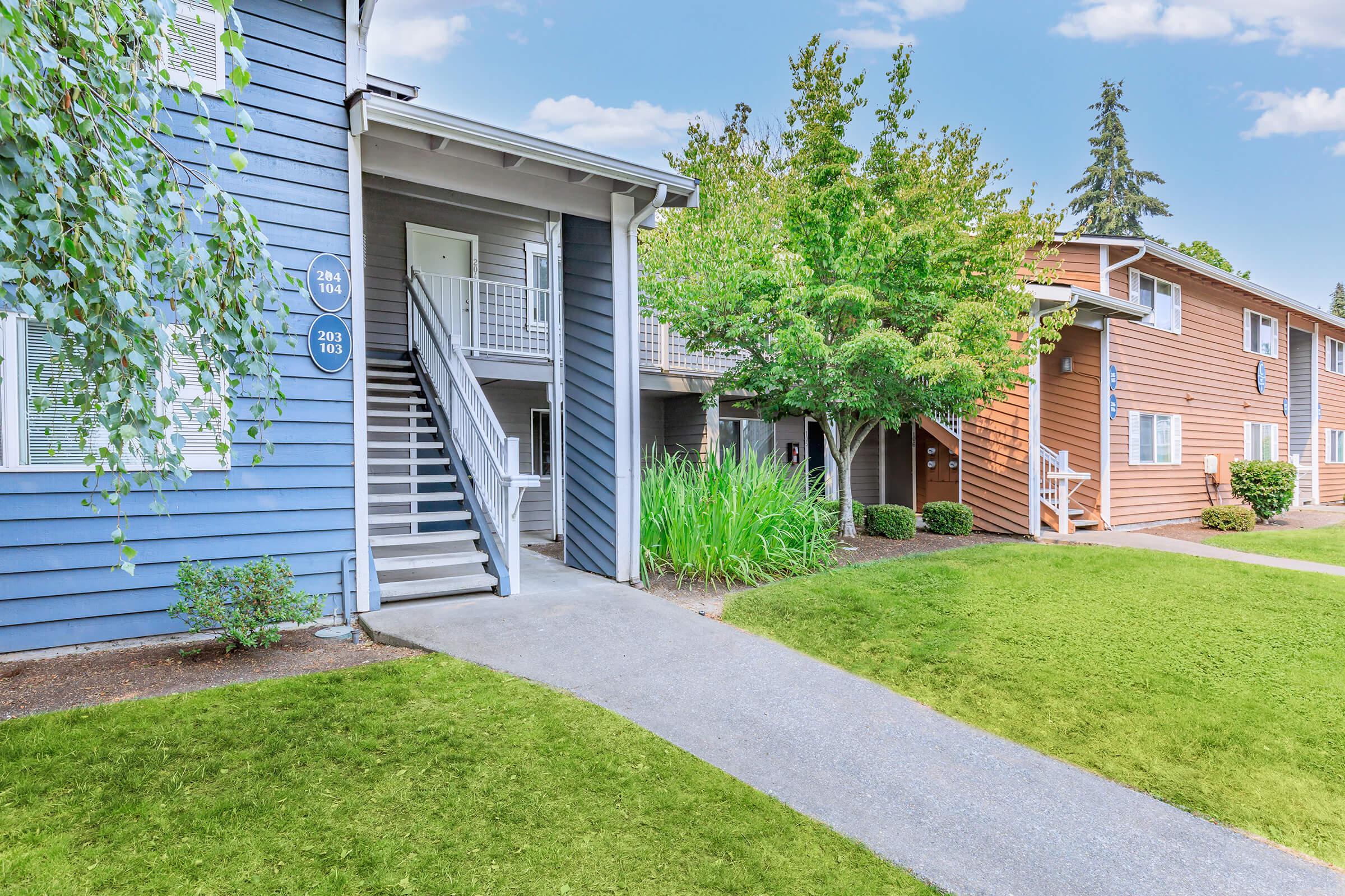 A serene apartment complex featuring two buildings, one blue and one warm-toned wood, with well-maintained landscaping. A pathway leads to the entrance of the blue building, flanked by greenery and shrubs. The scene is bright and inviting, set against a clear blue sky.