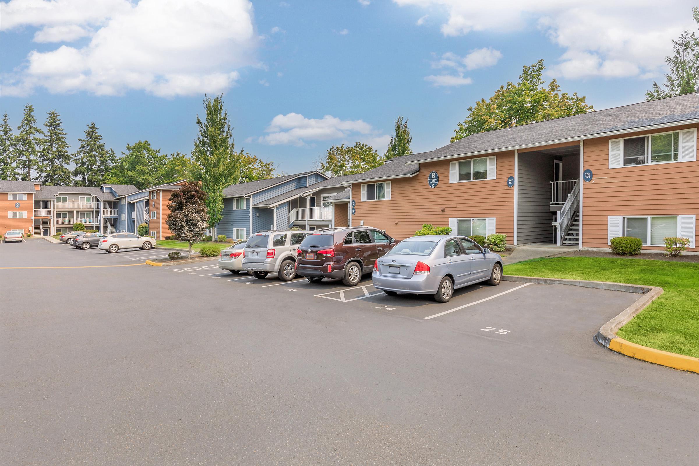A view of an apartment complex with two-story buildings featuring brown and blue exteriors. Several parked cars are in the foreground, with a clear sky and trees in the background. The scene is well-maintained, with green lawns and a paved driveway. The atmosphere is quiet and residential.