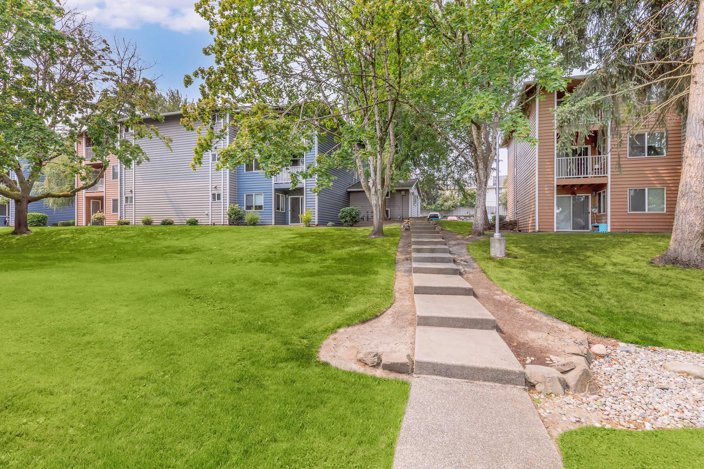 A well-maintained park-like area featuring a grassy lawn, trees, and two apartment buildings in the background. A concrete pathway winds through the green space, leading towards the entrances of the buildings. The scene is bright and inviting, with a clear sky above.