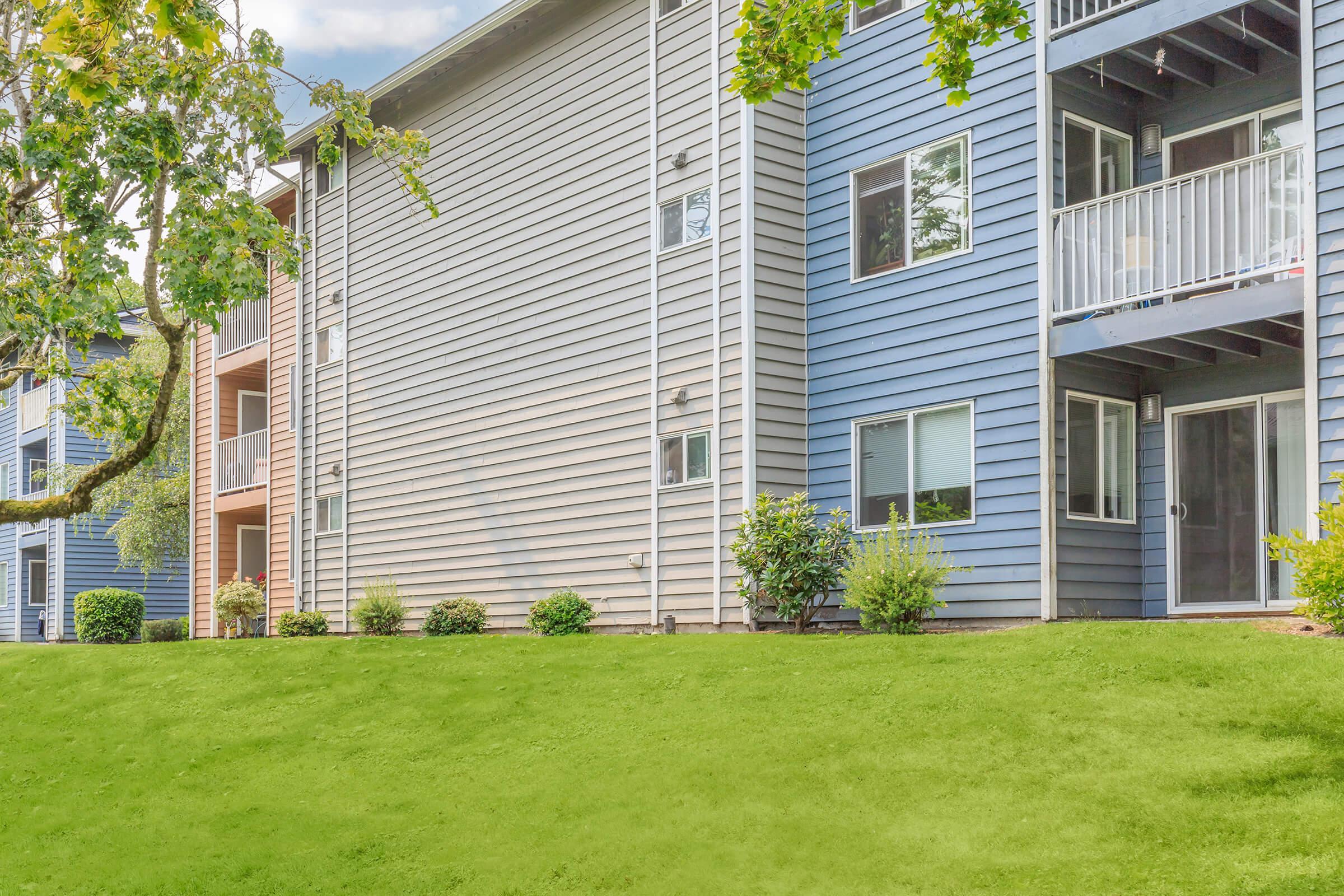 A row of apartment buildings with a mix of colors, featuring balconies and large windows. The grassy area in front is well-maintained with small shrubs. Trees can be seen on the left, adding greenery to the scene. The sky is partly cloudy, creating a pleasant atmosphere.