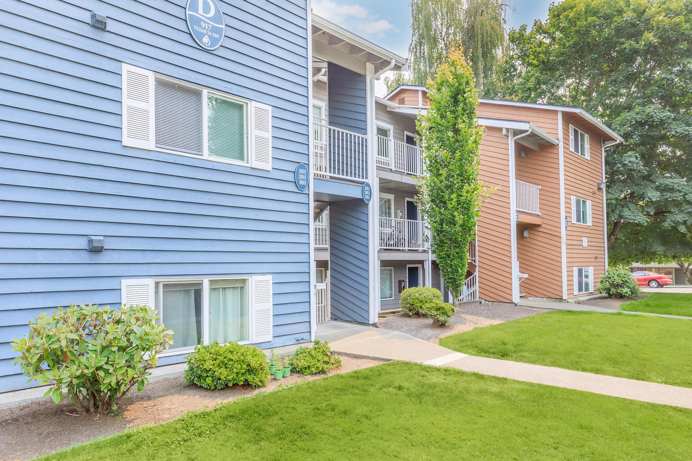 Two multi-story apartment buildings are shown, one painted light blue with white trim and balconies, and the other in brown. A well-manicured lawn with shrubs and a concrete pathway leads to the entrance. Trees are visible in the background, creating a serene residential environment.