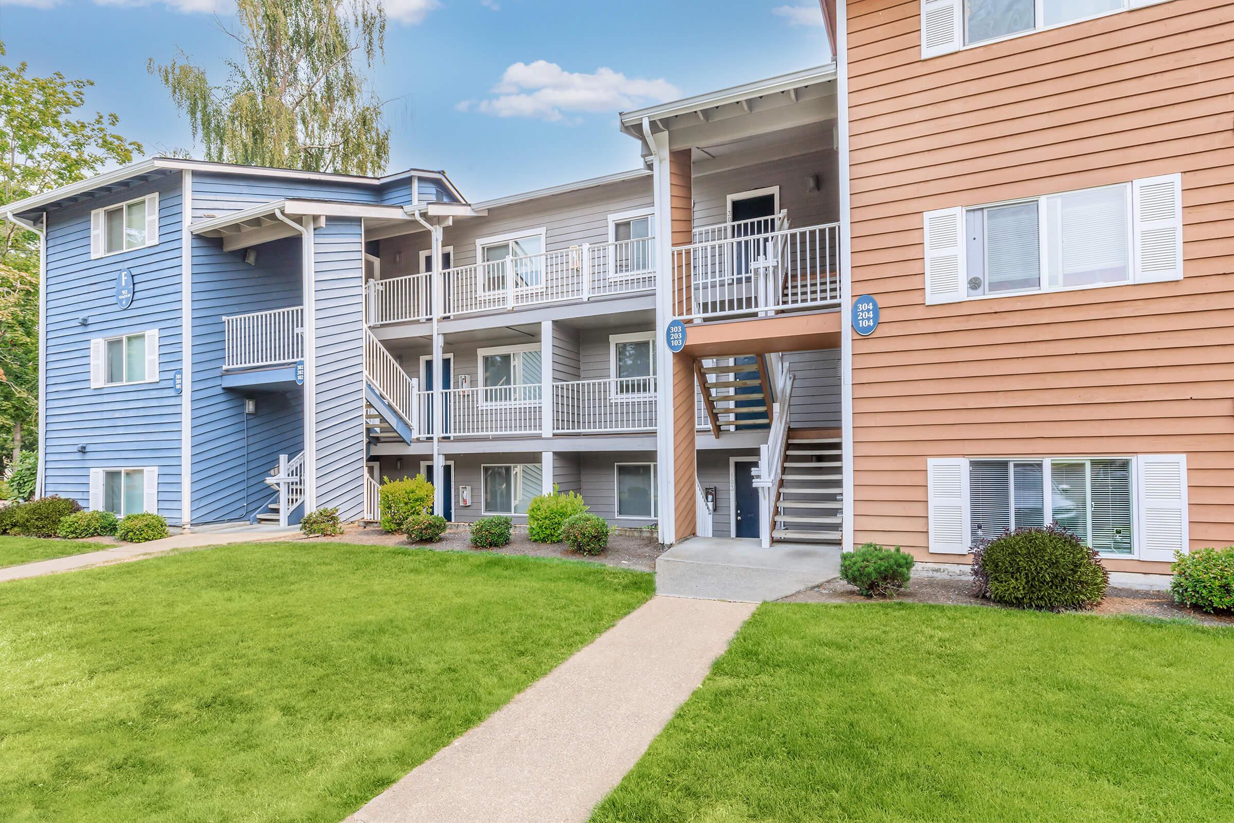 Two apartment buildings with blue and orange exteriors, featuring multiple balconies and white railings. The ground is landscaped with green grass and small bushes, and there is a concrete pathway leading to the entrance of the buildings. A clear blue sky is visible in the background.