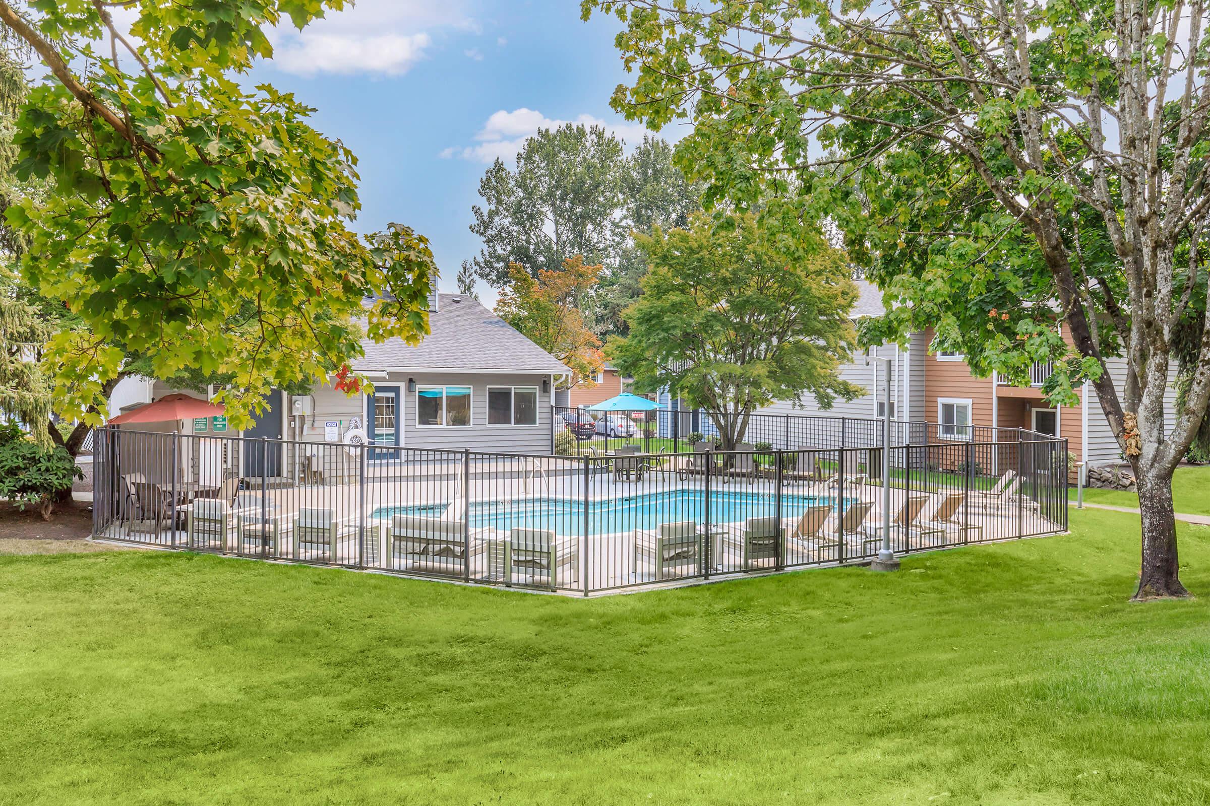 A fenced swimming pool surrounded by green grass and trees. Several lounge chairs are arranged around the pool, and colorful umbrellas provide shade. In the background, there are residential buildings and a clear blue sky.