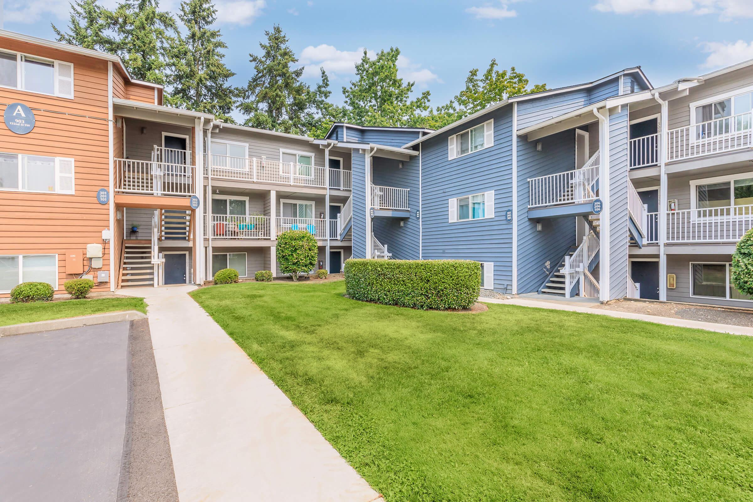 A view of a multi-unit apartment complex featuring two buildings in shades of orange and blue. The well-maintained grounds include a green lawn, bushes, and a walkway leading to the entrances of the units. The exterior has balconies and decorative signs near the doorways, set against a backdrop of trees and a blue sky.