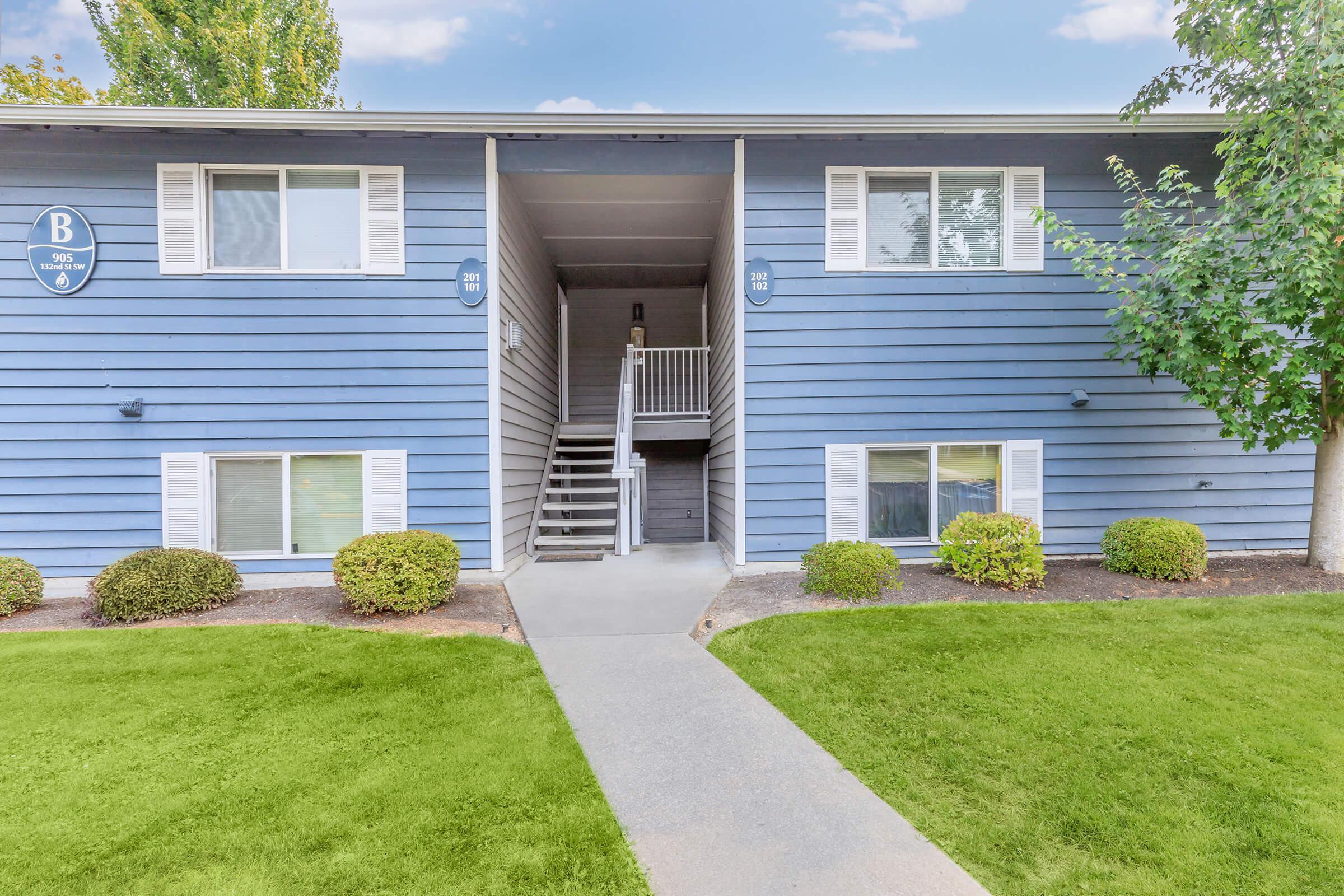 Exterior view of a two-story blue apartment building with white trim, featuring a central entrance and stairs leading to the upper level. The landscape includes neatly trimmed grass and small shrubs along the pathway. Clear blue sky and trees in the background enhance the inviting atmosphere.