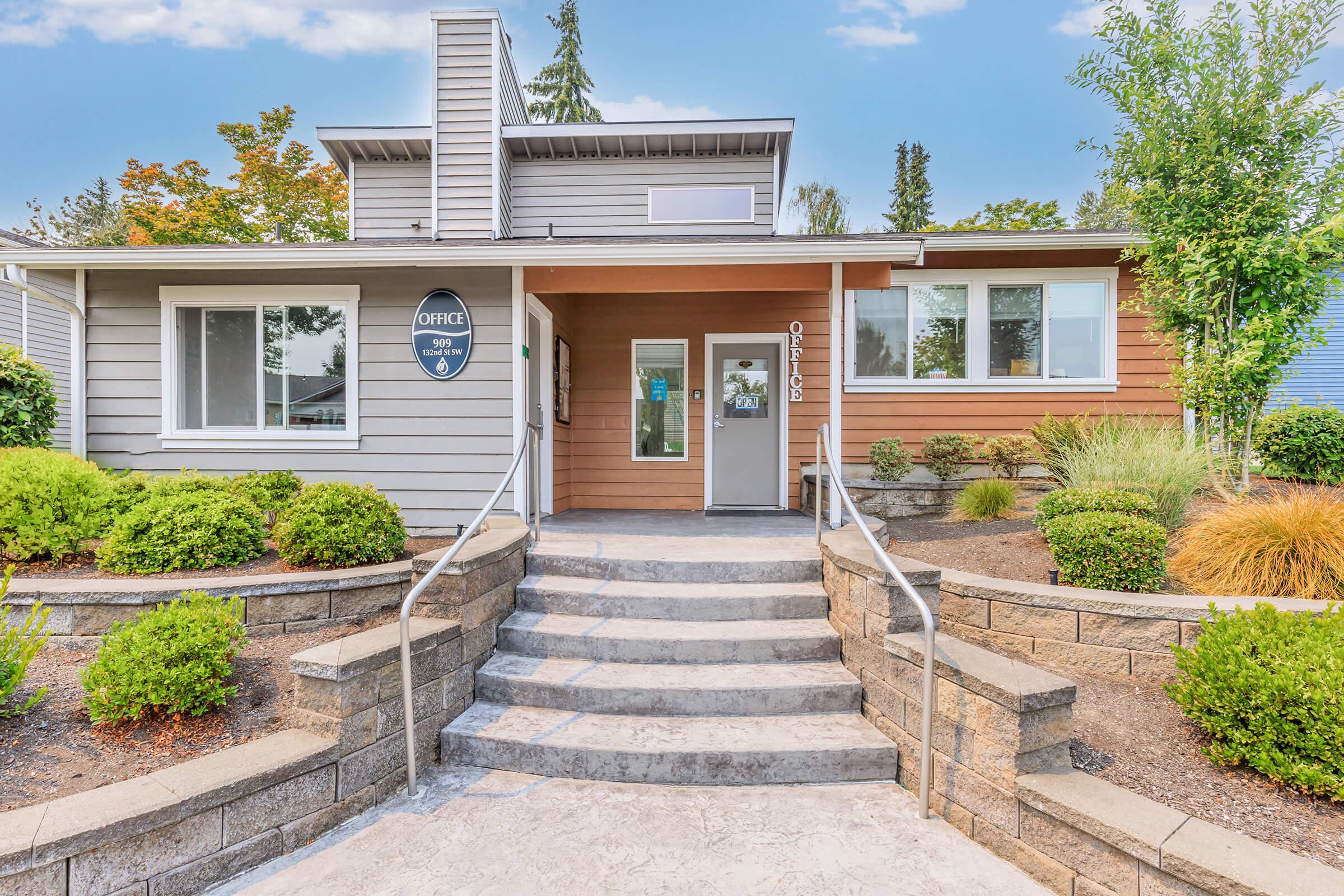 A modern office building with a welcoming entrance, featuring a set of stairs leading up to a wooden door, surrounded by neatly landscaped greenery and shrubs. The exterior combines gray siding with wooden accents, and there are large windows allowing natural light into the space.
