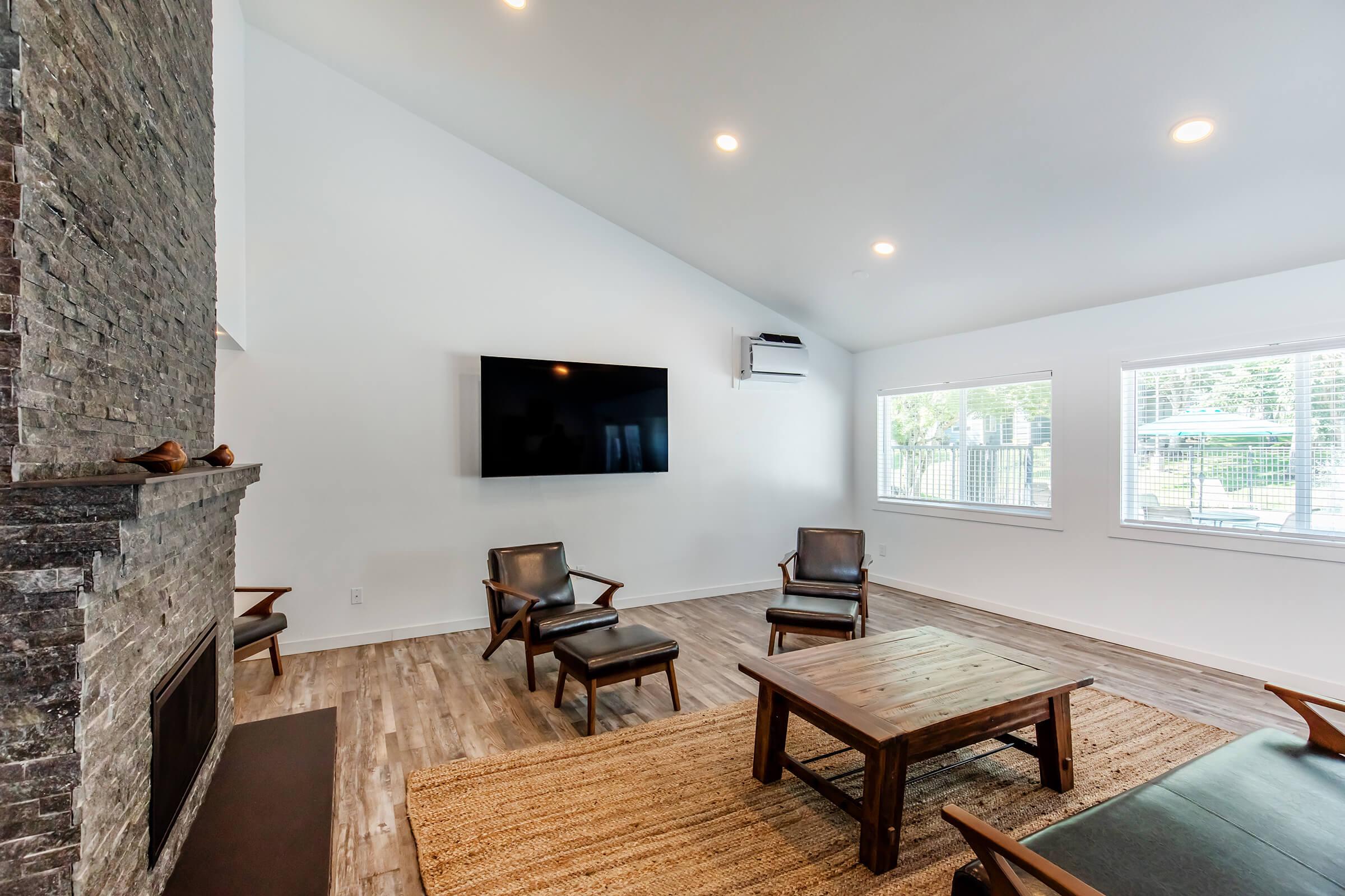 A modern living room featuring a high ceiling, hardwood floors, and a stone fireplace. There are two stylish wooden chairs and a low wooden coffee table on a natural fiber rug. A flat-screen TV is mounted on the wall, and large windows allow natural light to fill the space.