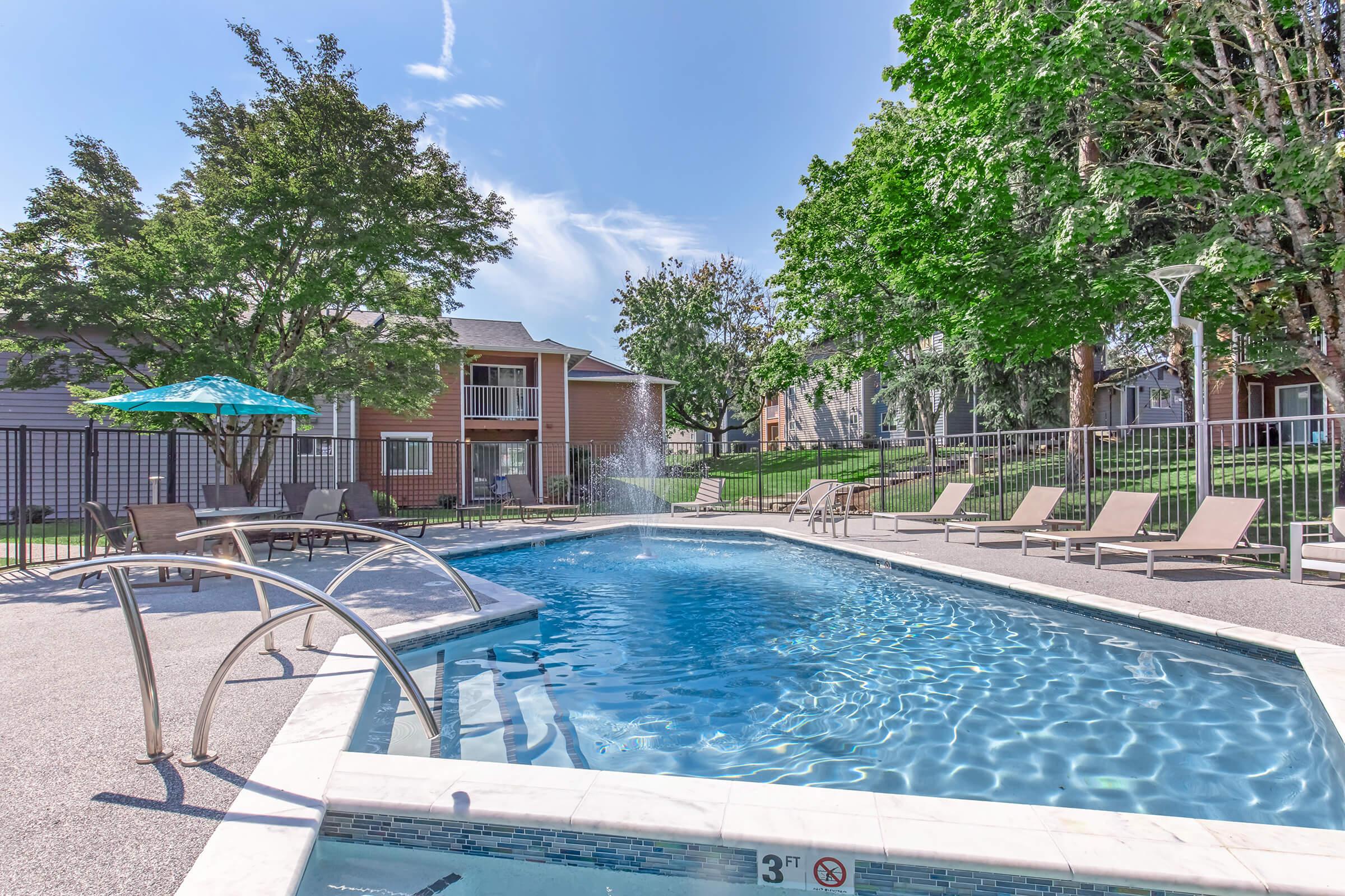 A crystal-clear swimming pool surrounded by lounge chairs and an umbrella. Green trees provide shade in the background, while nearby buildings add to the serene atmosphere. A fountain sprays water in the center of the pool, enhancing the inviting environment.