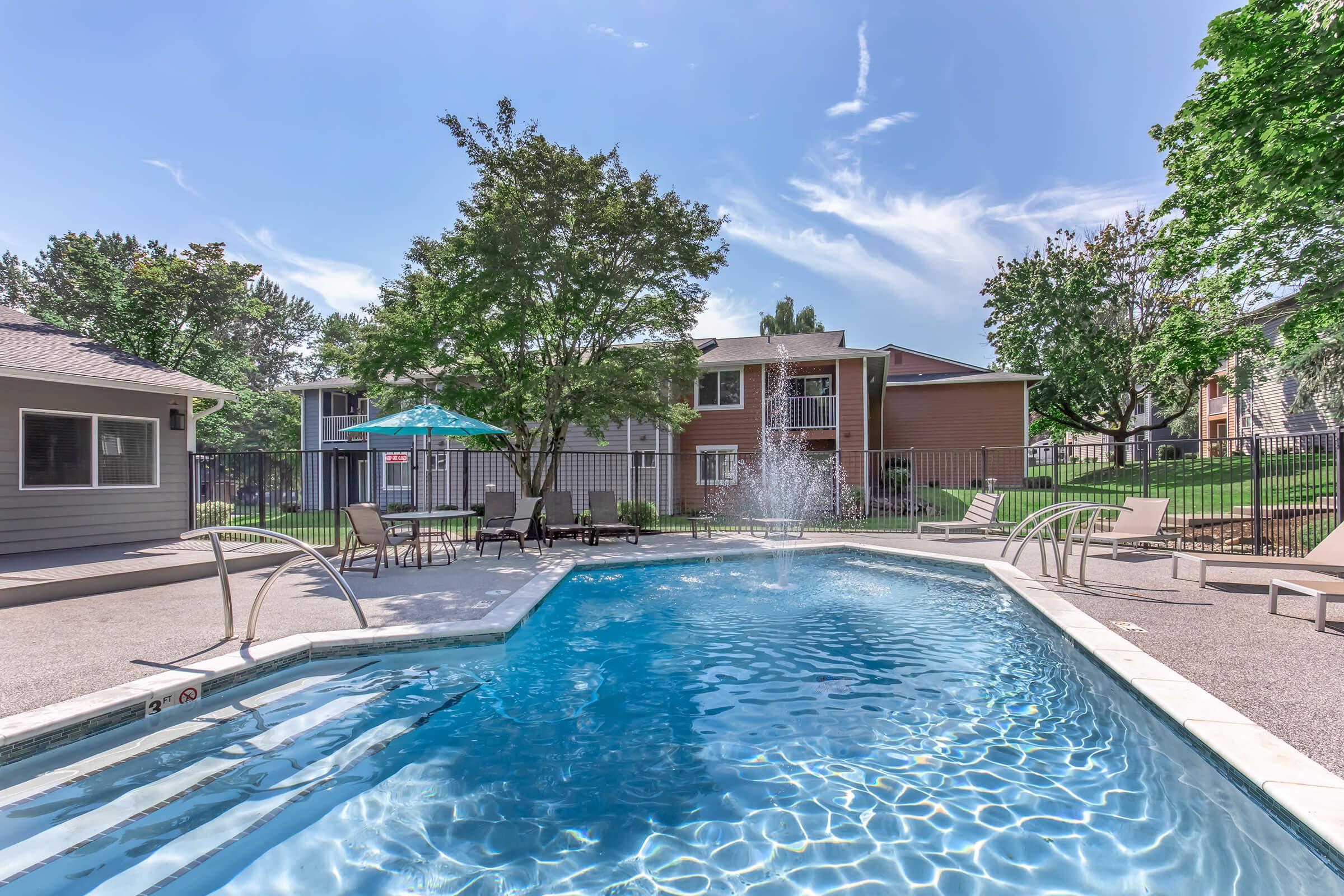 A clear blue swimming pool surrounded by lounge chairs, greenery, and trees. Two apartment buildings are visible in the background, and a colorful umbrella provides shade near the pool area. The scene is bright and sunny, creating a relaxing outdoor atmosphere.