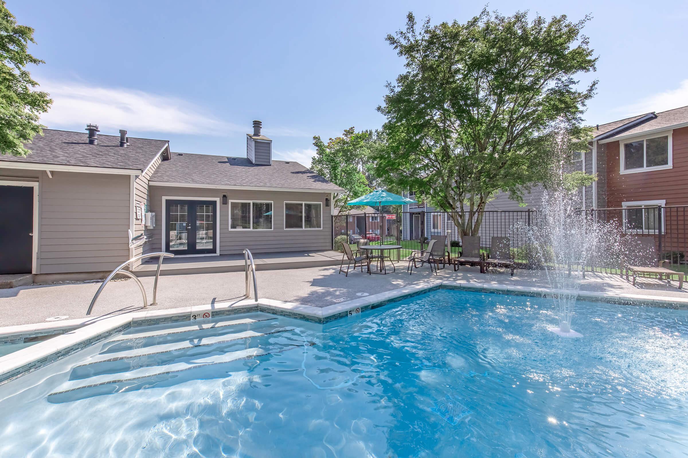 A clear swimming pool with a fountain feature, surrounded by lounge chairs and shaded tables. In the background, there are two buildings with varied colors, well-maintained landscaping, and a sunny sky. The setting suggests a relaxing outdoor area, ideal for leisure and social gatherings.