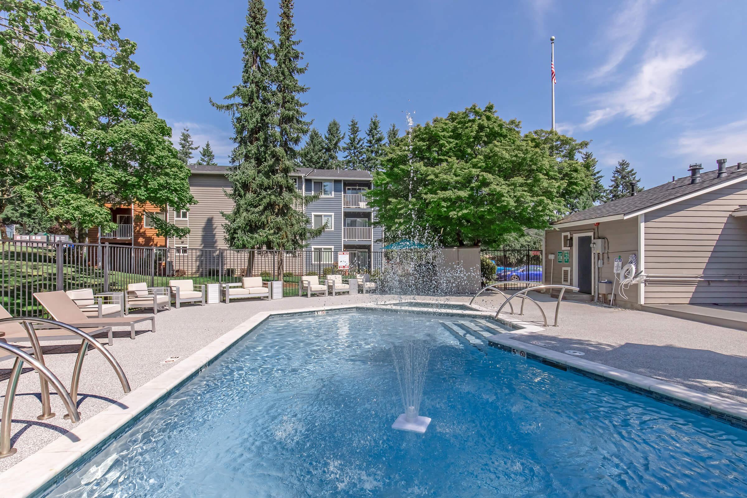 A clear swimming pool with a fountain feature, surrounded by lounge chairs, greenery, and a residential building in the background. Trees provide shade, and the sky is blue with a few clouds. An American flag is visible in the distance.