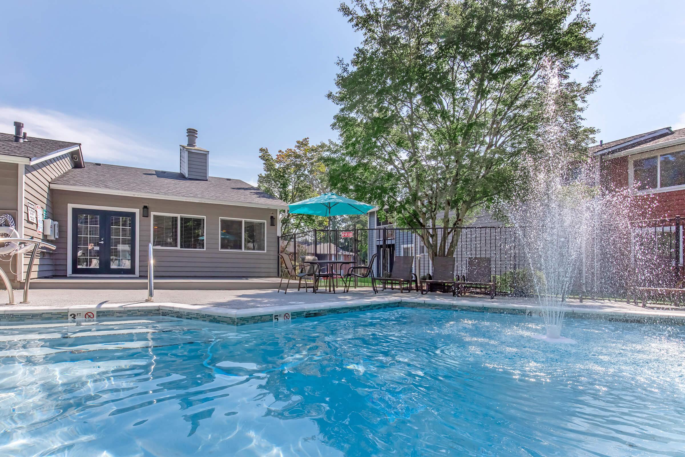 A serene swimming pool scene featuring crystal-clear water and a fountain spray. In the background, there’s a cozy house with a patio, shaded by a large umbrella. Lush trees provide greenery, creating a relaxing outdoor atmosphere with lounge chairs and tables nearby.