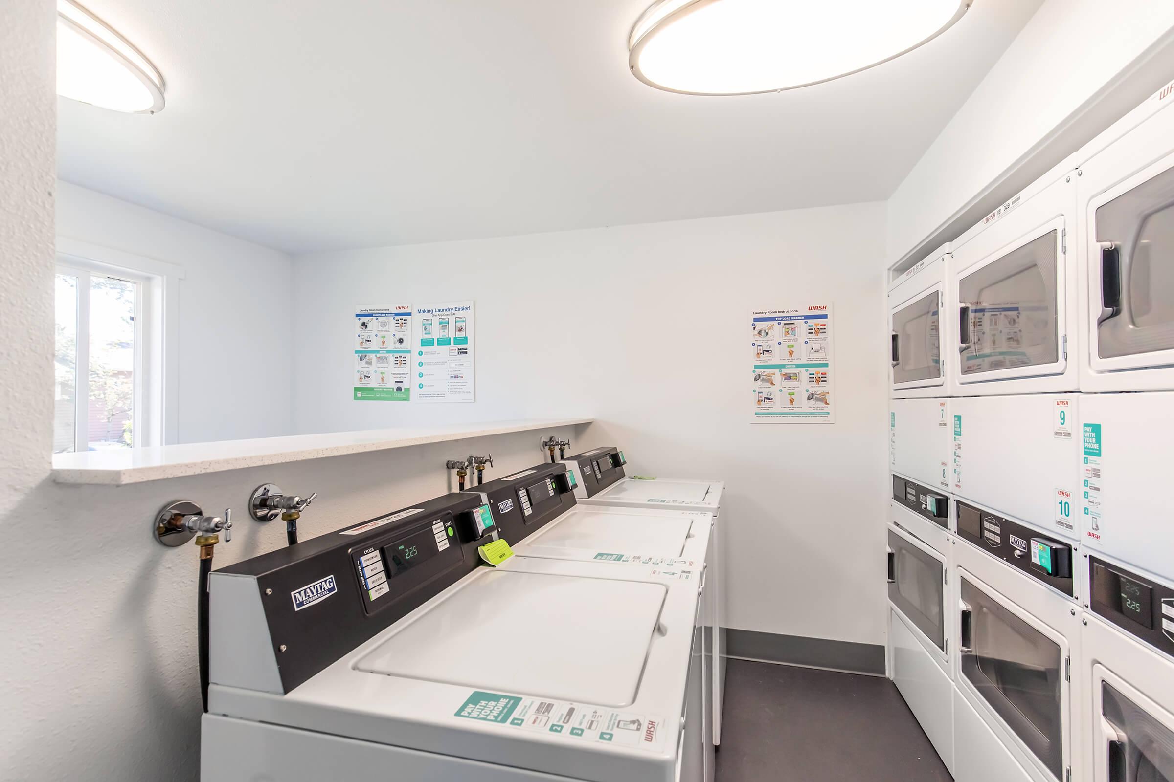 A clean, well-lit laundry room featuring multiple washing machines and dryers. The machines are neatly lined up against a white wall. There are informational posters on the wall, and natural light filtering in from a nearby window. The floor is gray, adding to the modern aesthetic.