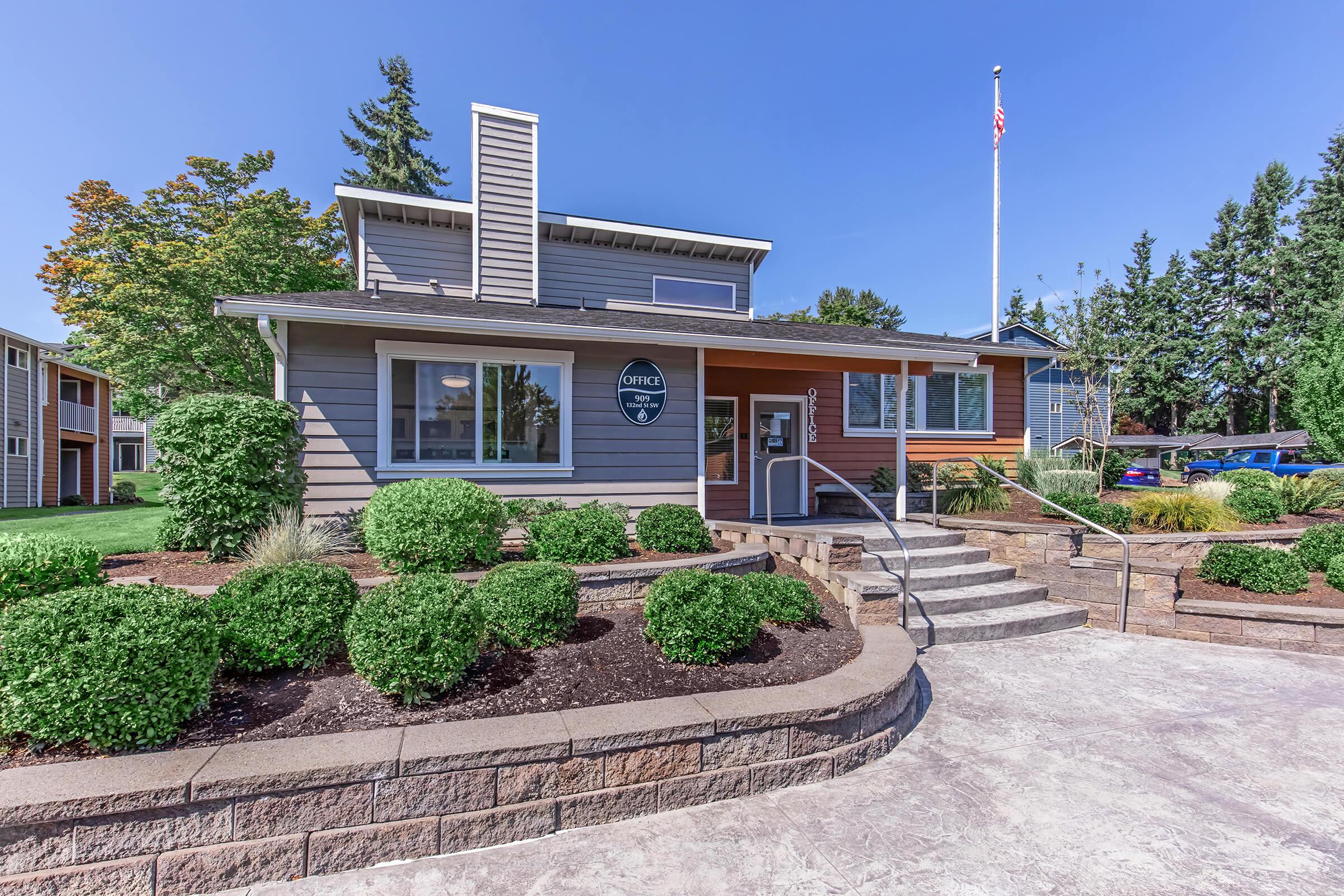A modern gray and wood-paneled house with landscaped green shrubs and stone steps leading to the entrance. The house features large windows and a flagpole with an American flag. The sky is clear and blue, complementing the well-maintained yard and neighboring homes in the background.