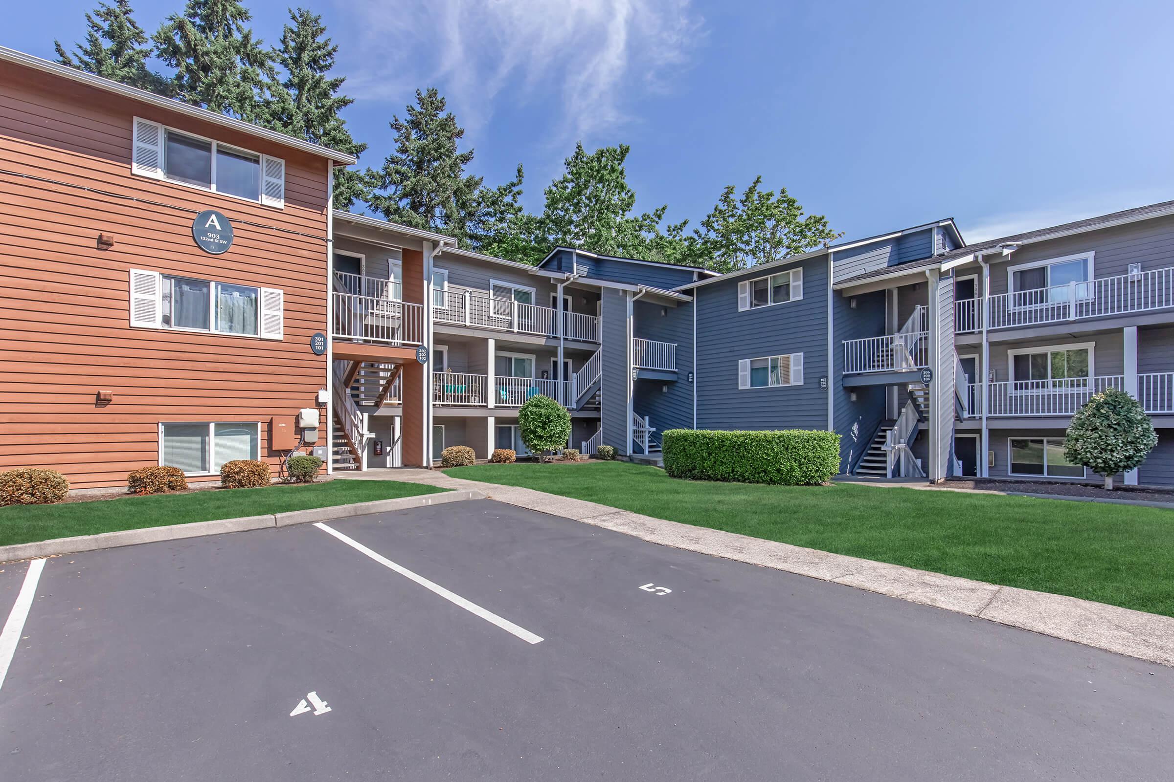 A multi-unit residential complex featuring two buildings, one in orange and one in blue, surrounded by landscaped grass. There are balconies on the upper floors, and parking spaces are visible in the foreground. The sky is clear with a few clouds.