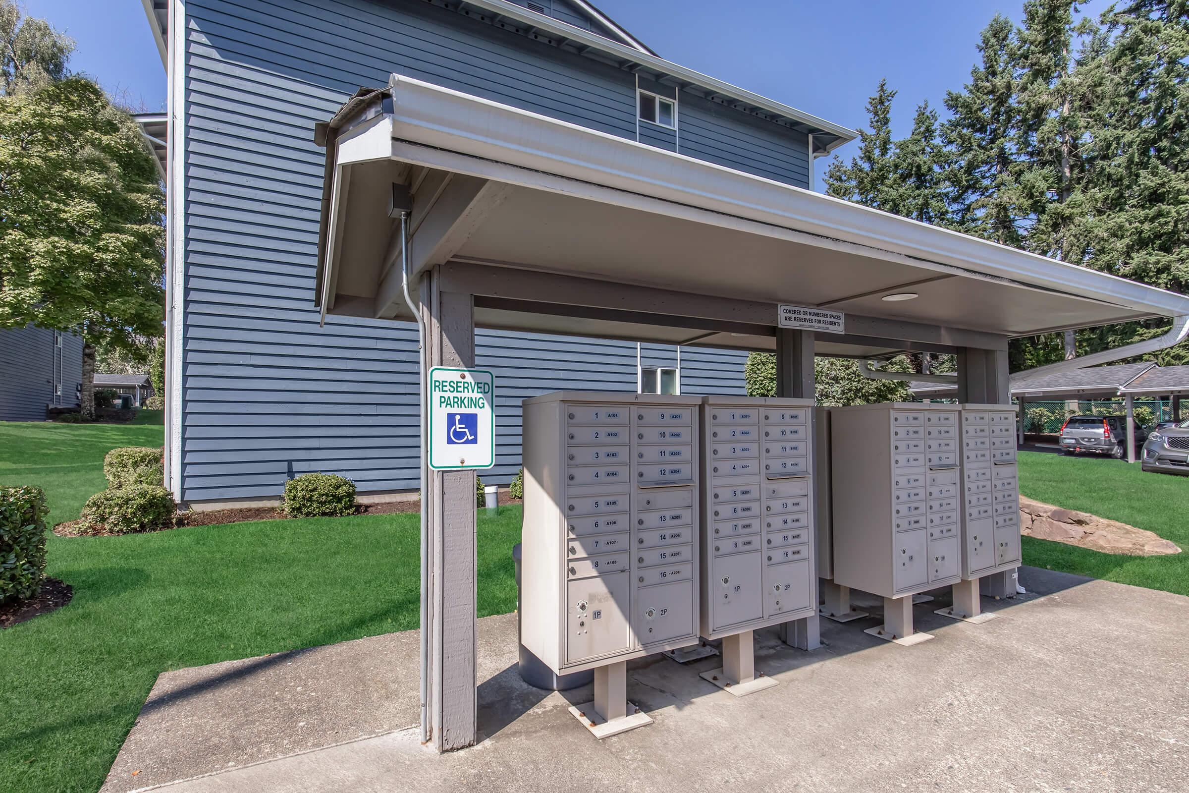 Covered mailbox area adjacent to a multi-unit building. The structure features multiple mailboxes and a sign indicating reserved parking, specifically for disabled individuals. Surrounding the area is well-maintained grass and shrubs, providing a neat and accessible environment.