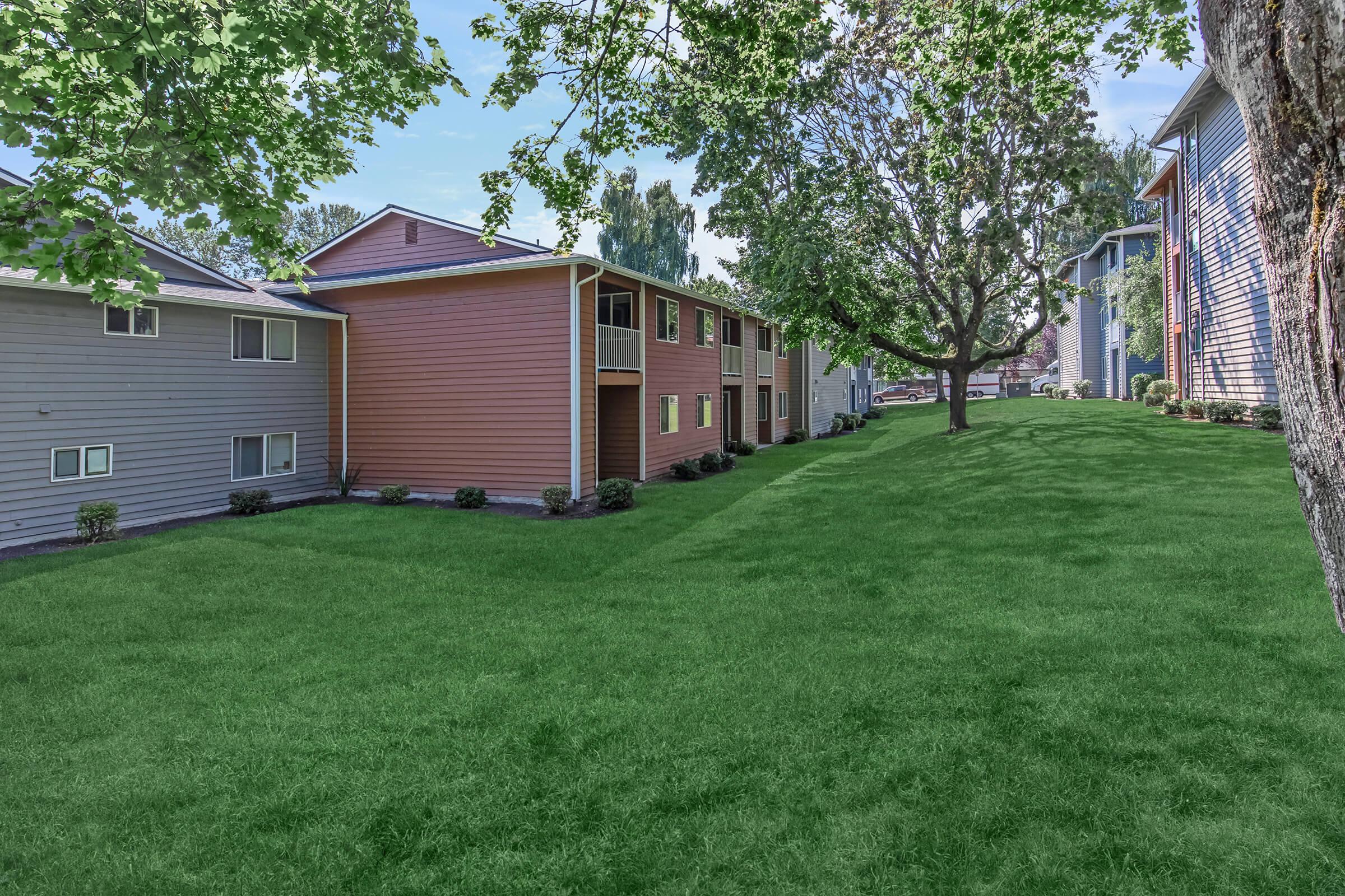 A grassy area between two rows of residential buildings with trees and landscaping. The buildings have varying colors, and the scene is bright and sunny, suggesting a well-maintained outdoor space.