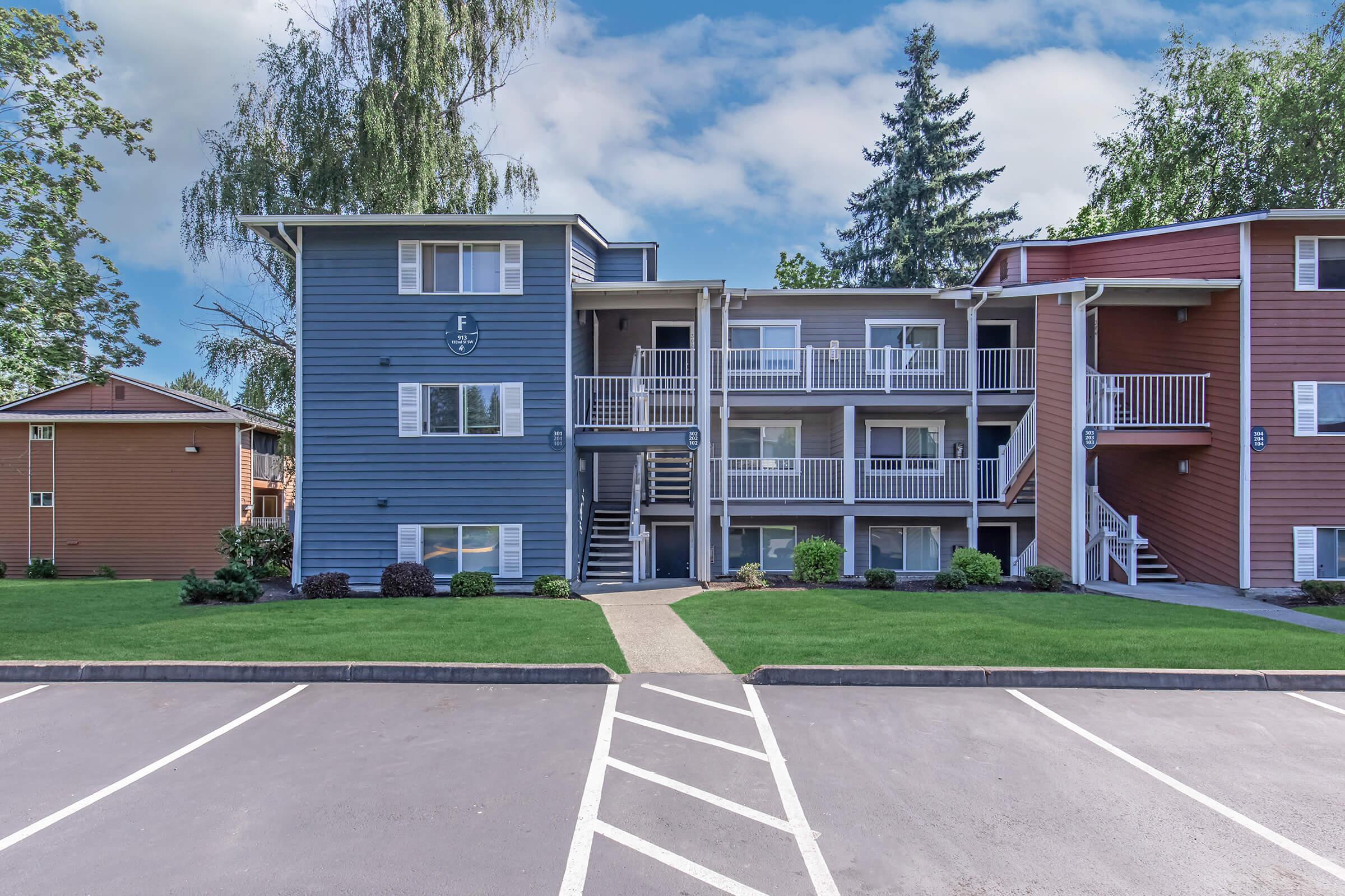 A two-story apartment building with a mix of blue and brown siding. It features outdoor staircases, balconies, and well-maintained landscaping with green grass and trees. A parking lot is visible in the foreground with clearly marked parking spaces. The sky is partly cloudy.