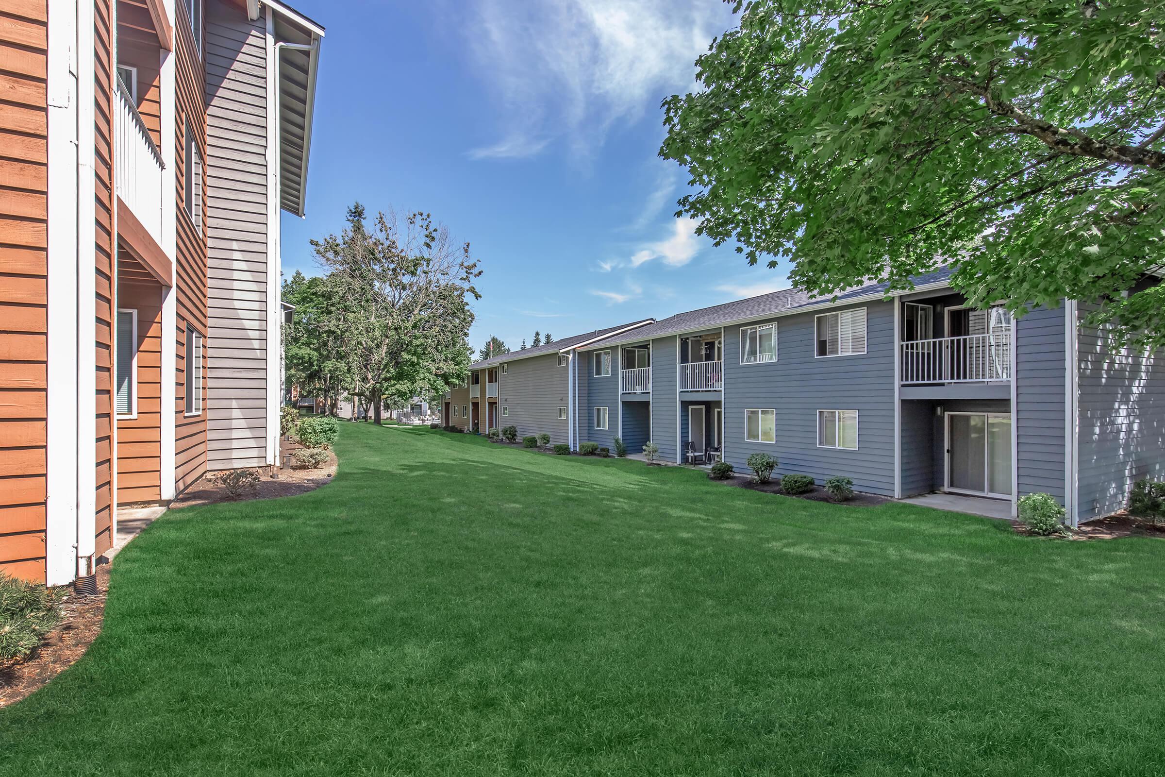 A view of a landscaped area between two apartment buildings, one with orange siding and the other in light blue. The grassy space is lush and well-maintained, surrounded by small shrubs and trees under a blue sky with some clouds.