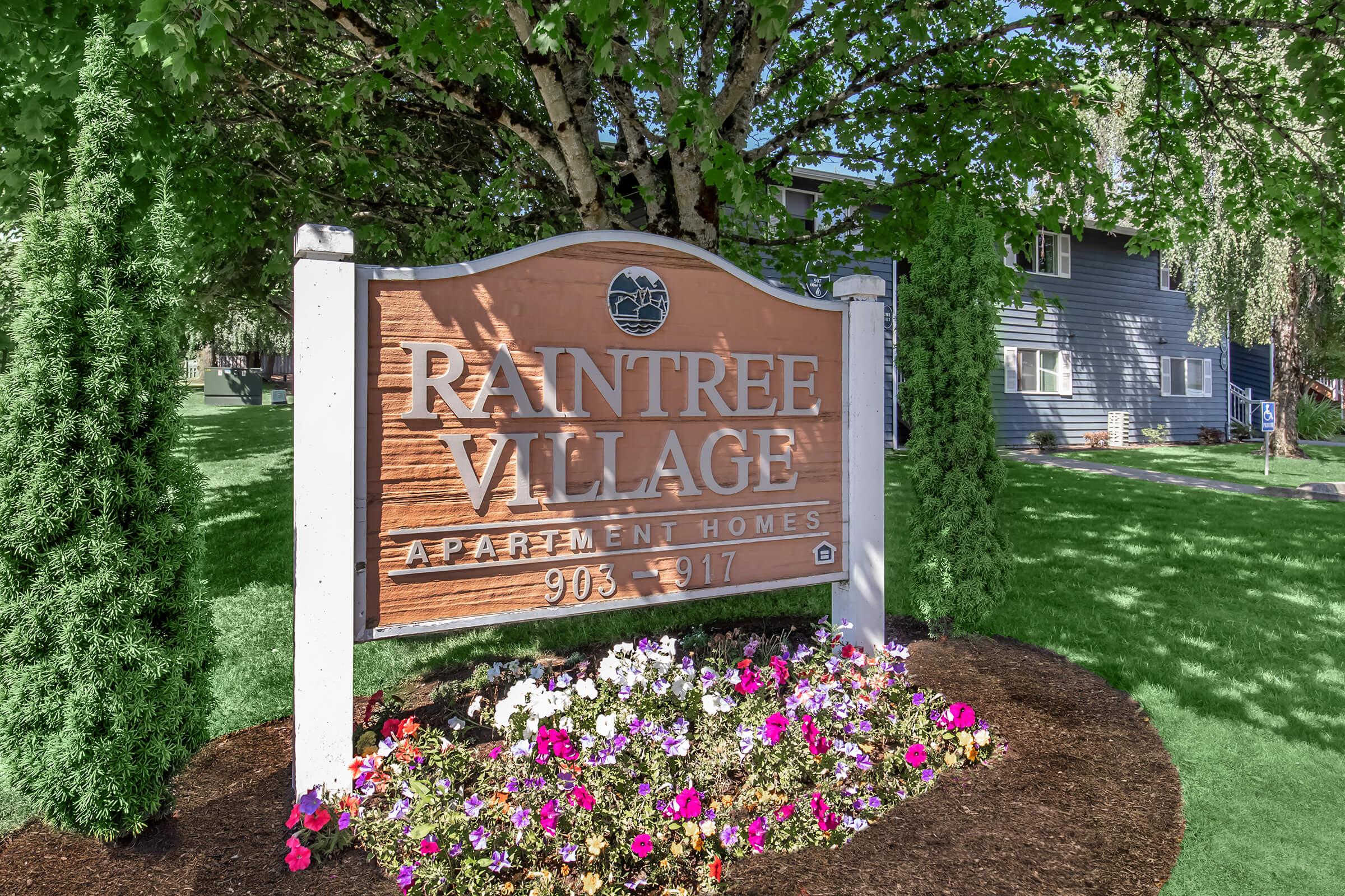 Sign for Raintree Village Apartment Homes, featuring the name prominently displayed on a wooden background. Surrounded by colorful flowers and greenery, the sign provides the address range of 905-917. The setting appears well-maintained and inviting, emphasizing a residential community atmosphere.
