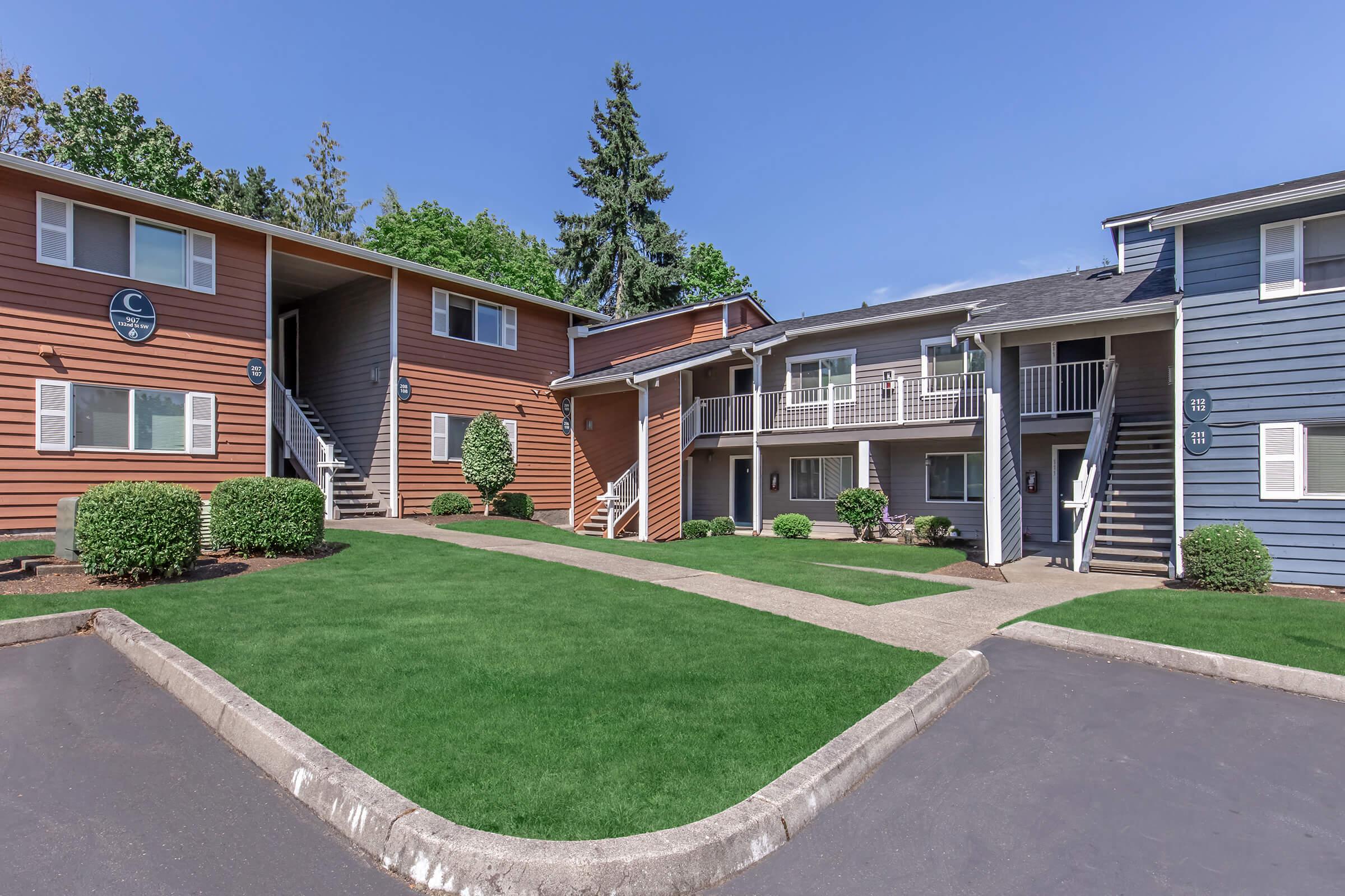 Two multi-unit residential buildings in a well-maintained complex. One building is painted orange, while the other is in blue. A short pathway leads between them, and the landscaping features manicured green lawns and small bushes. The sky is clear and sunny, creating a vibrant atmosphere.
