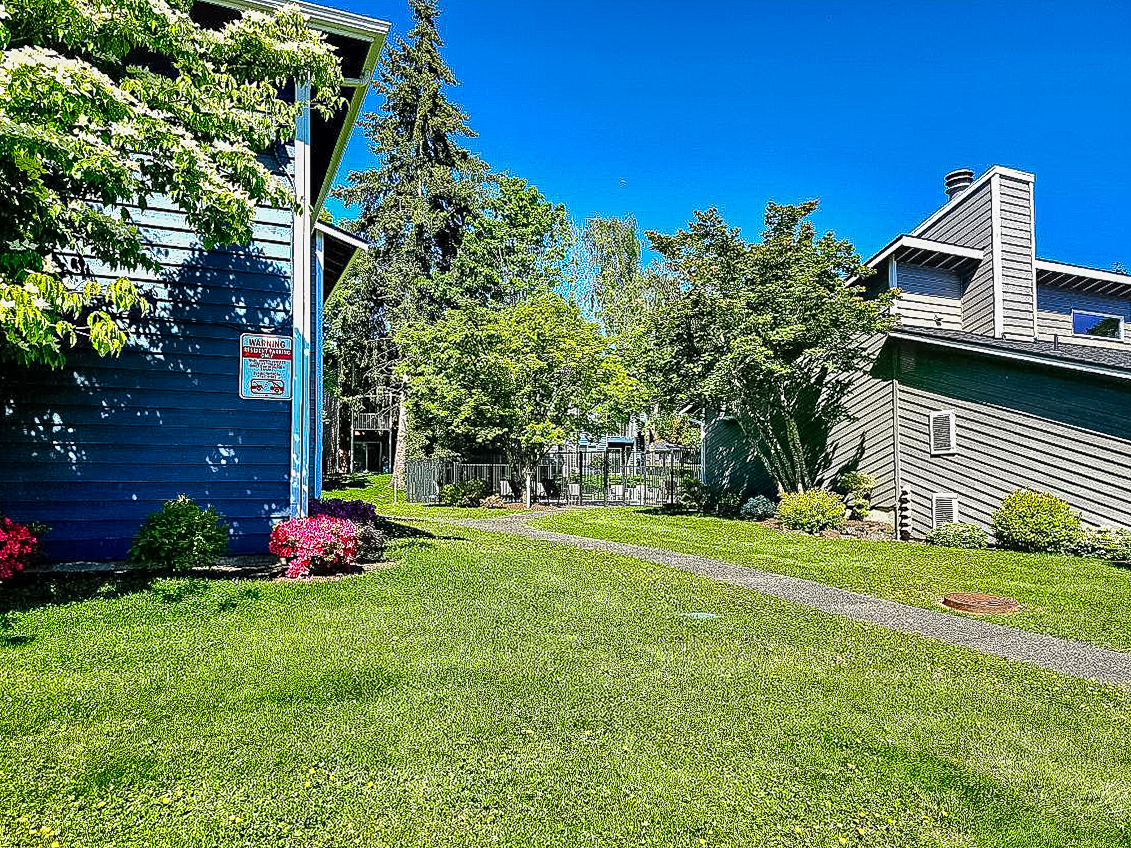 A bright, sunny outdoor scene featuring a well-maintained pathway lined with grass and shrubs, flanked by two houses, one blue and one gray. Trees and greenery create a lush environment, with vibrant flowers blooming near the pathway. Clear blue sky overhead enhances the cheerful atmosphere.