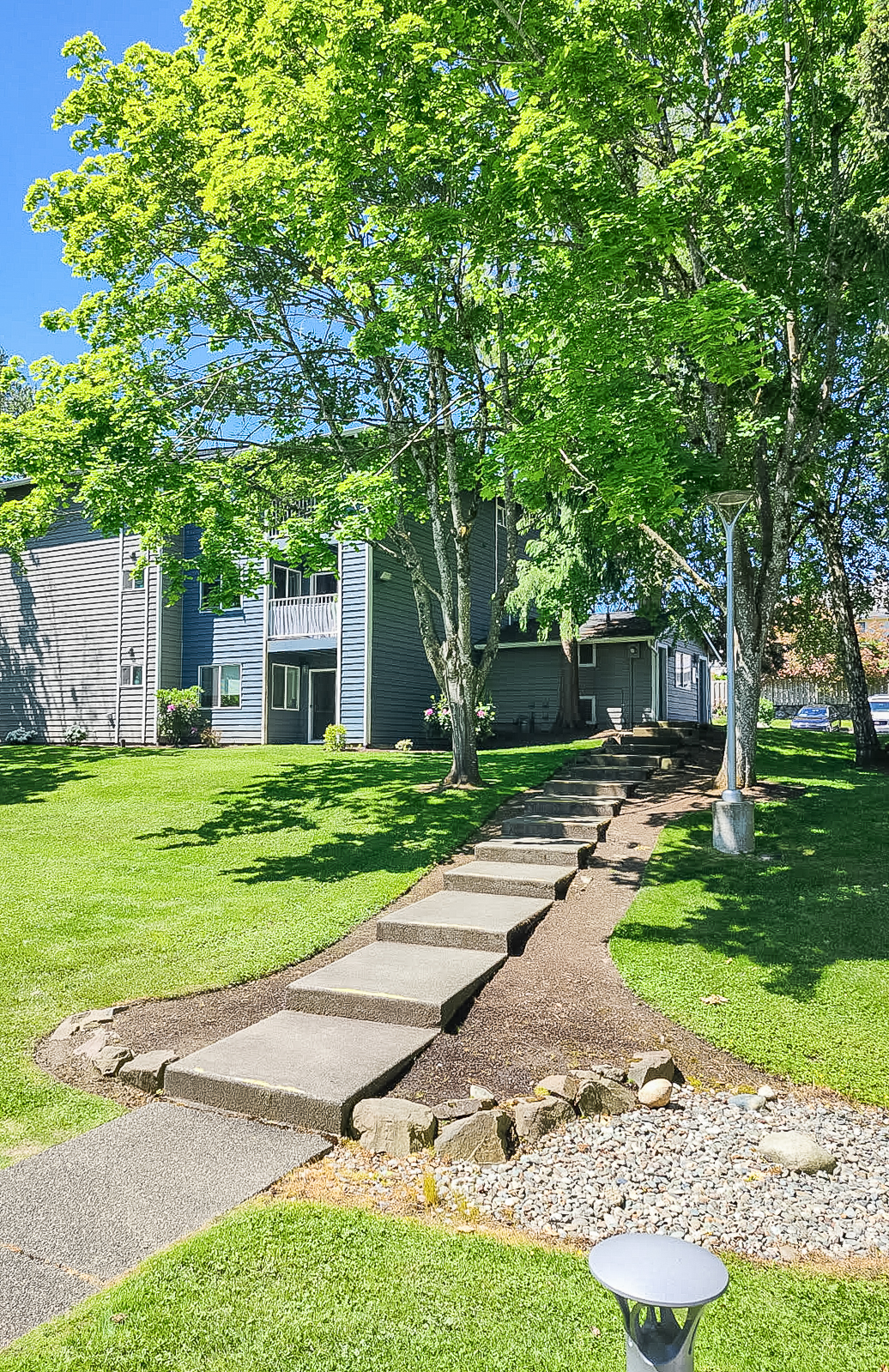 A pathway of stepping stones lined with gravel leads through a green lawn to a multi-story blue building surrounded by trees. The scene is bright and sunny with clear blue skies, showcasing a peaceful and well-maintained outdoor environment.