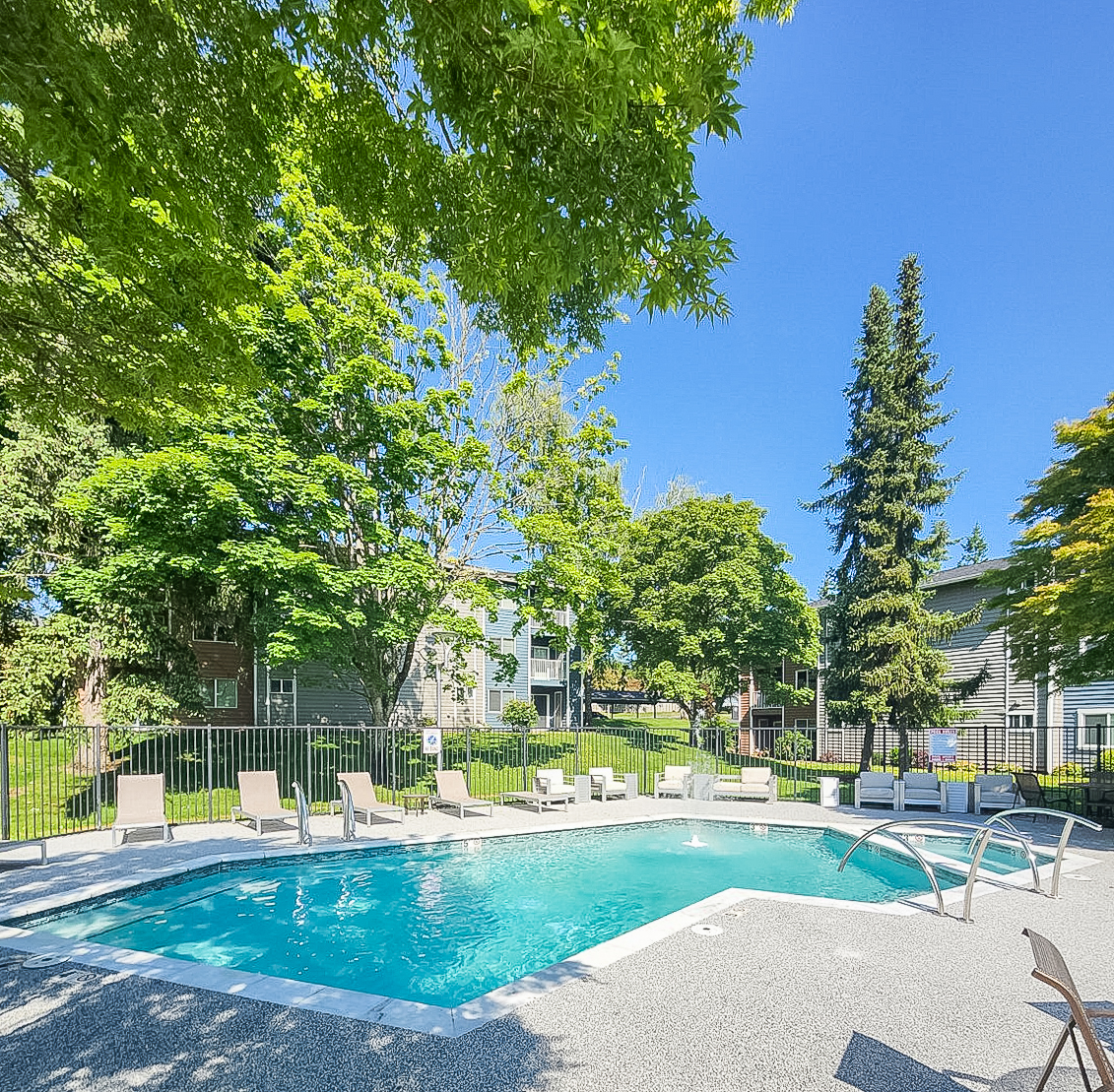 A sunny outdoor pool area surrounded by greenery and residential buildings. The pool is clear and inviting, with lounge chairs set up around it. Tall trees provide shade, and the sky is bright blue, creating a relaxing atmosphere for swimming and lounging.