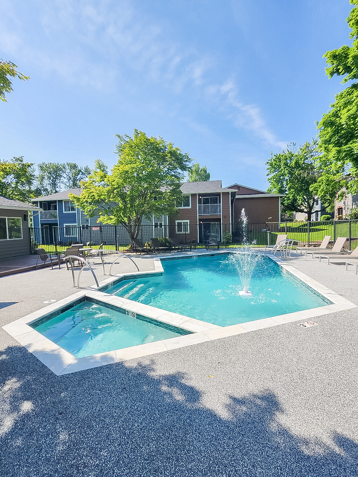 A sunny outdoor pool area featuring a blue swimming pool with a fountain, surrounded by lounge chairs on a gravel surface. In the background, there are green trees and residential buildings, with a clear blue sky overhead, creating a relaxing summer atmosphere.
