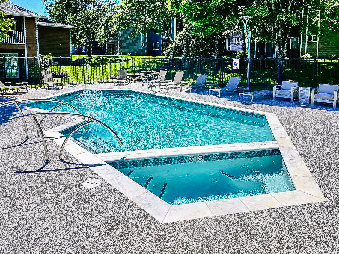 A clear swimming pool with a unique shape, surrounded by lounge chairs on a textured surface. There are green trees and buildings in the background, and a gated area for safety. The pool features a shallow end marked with "3 ft." signage for depth indication. Sunlight reflects off the water, creating a bright and inviting atmosphere.