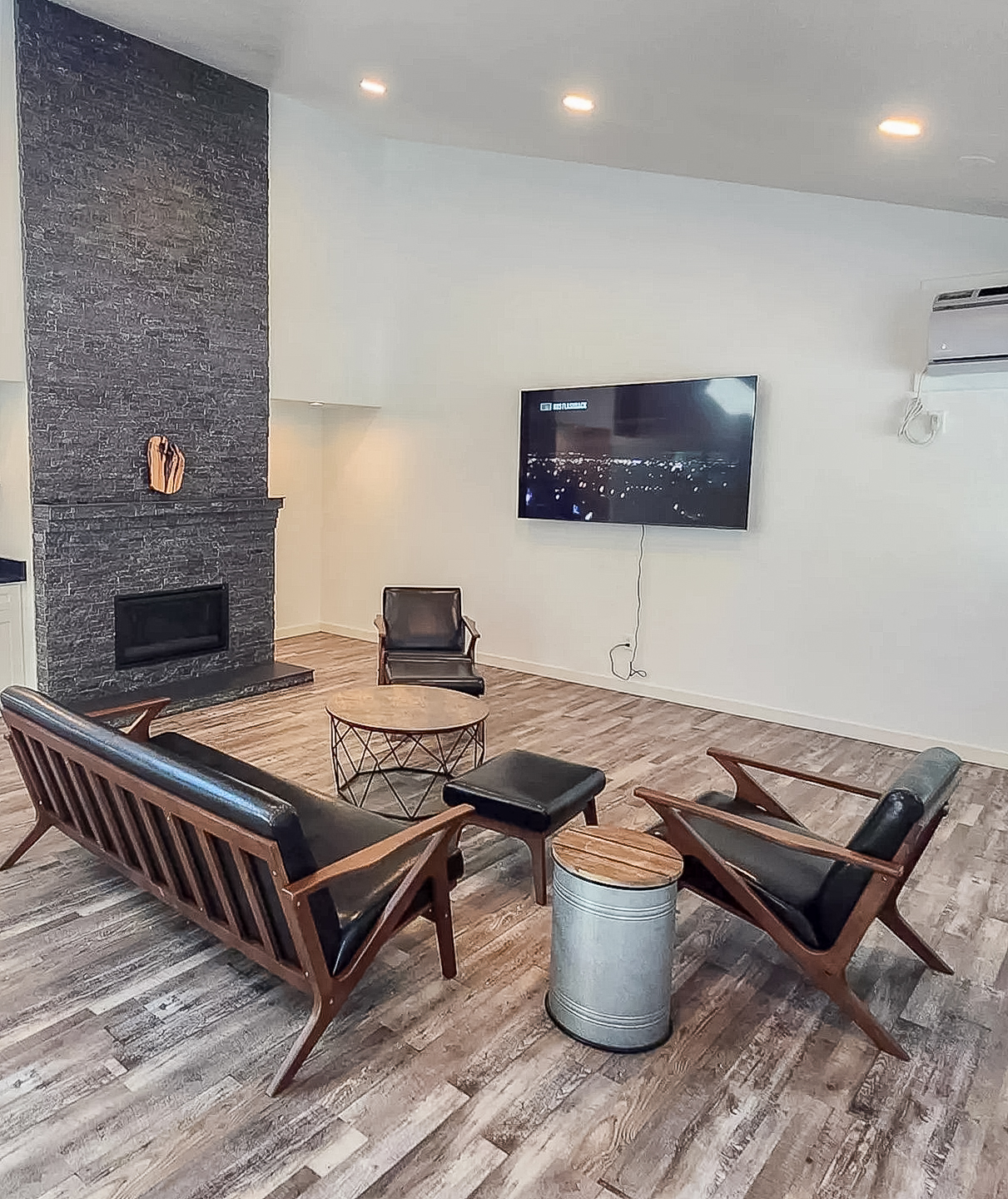 A modern living room featuring a stone fireplace, a wall-mounted TV, and stylish furniture. The seating includes two sleek wooden chairs, a circular coffee table, and a small metallic side table. The space has a light ambiance with wooden flooring and minimal decor, creating a cozy and inviting atmosphere.