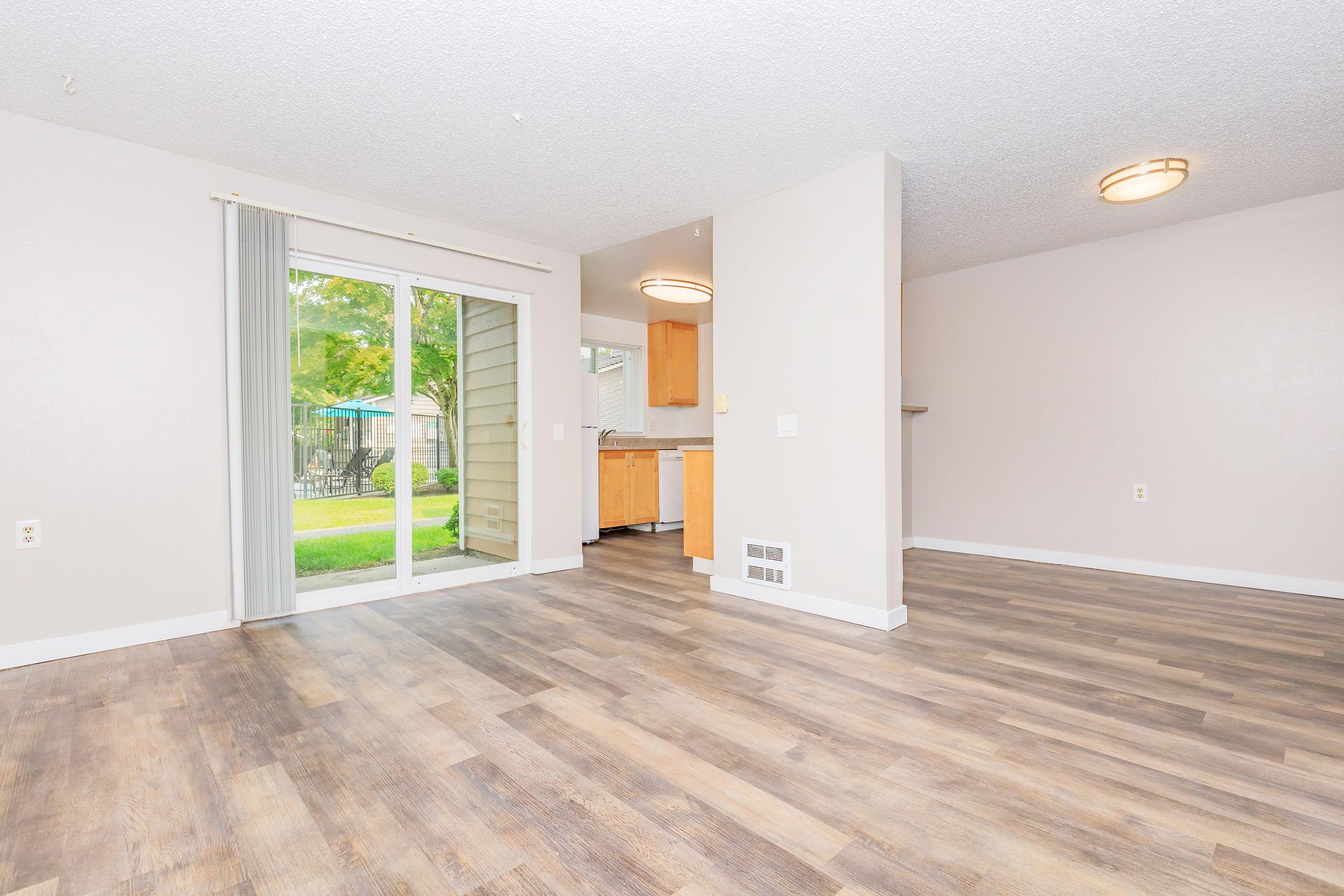 Spacious living area with light-colored walls and wood-style flooring. Sliding glass doors lead to an outdoor space with greenery. A kitchen is visible in the background, featuring wooden cabinetry. The room is well-lit with natural light from the outside. Suitable for a cozy living or dining setup.