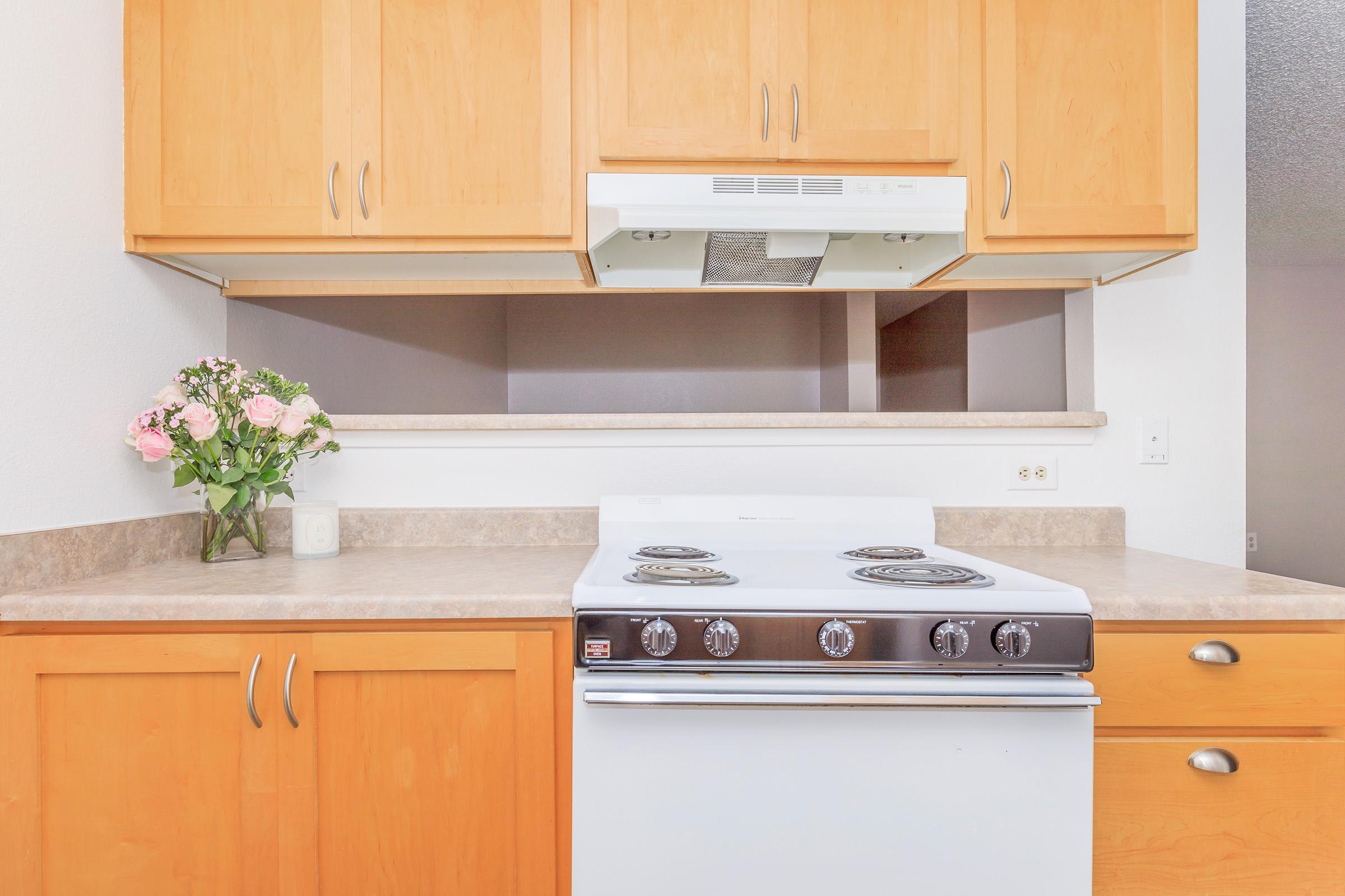 A clean and modern kitchen featuring light wood cabinetry, a white stove with four burners, and a ventilation hood. On the countertop, there's a small white candle and a vase of pink roses, adding a touch of elegance to the space. The overall aesthetic is bright and inviting.