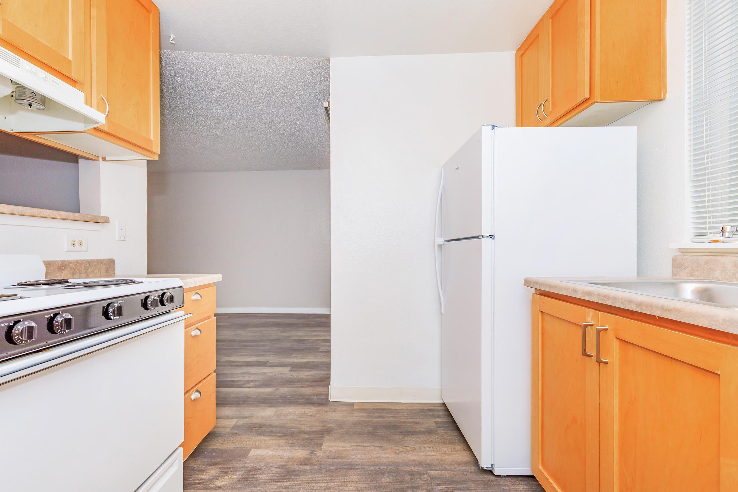 A bright kitchen featuring wooden cabinets, a white refrigerator, a gas stove with four burners, and a sink. The flooring is a modern laminate, and there's a glimpse of a spacious living area in the background, with neutral-colored walls and ample natural light.