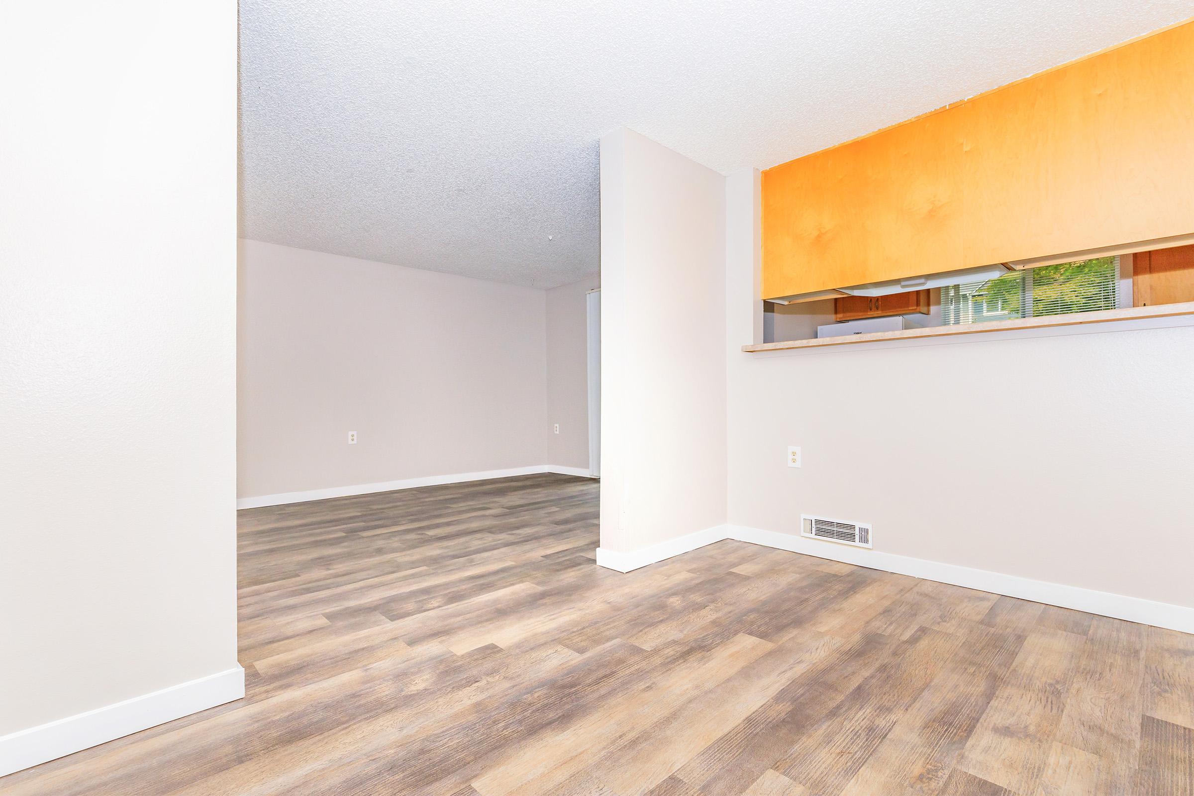 Spacious interior of a well-lit room featuring light wood-look vinyl flooring, neutral walls, and an open layout. There is a partial view of a kitchen area with a light orange backsplash. The space appears inviting and suitable for various design possibilities.