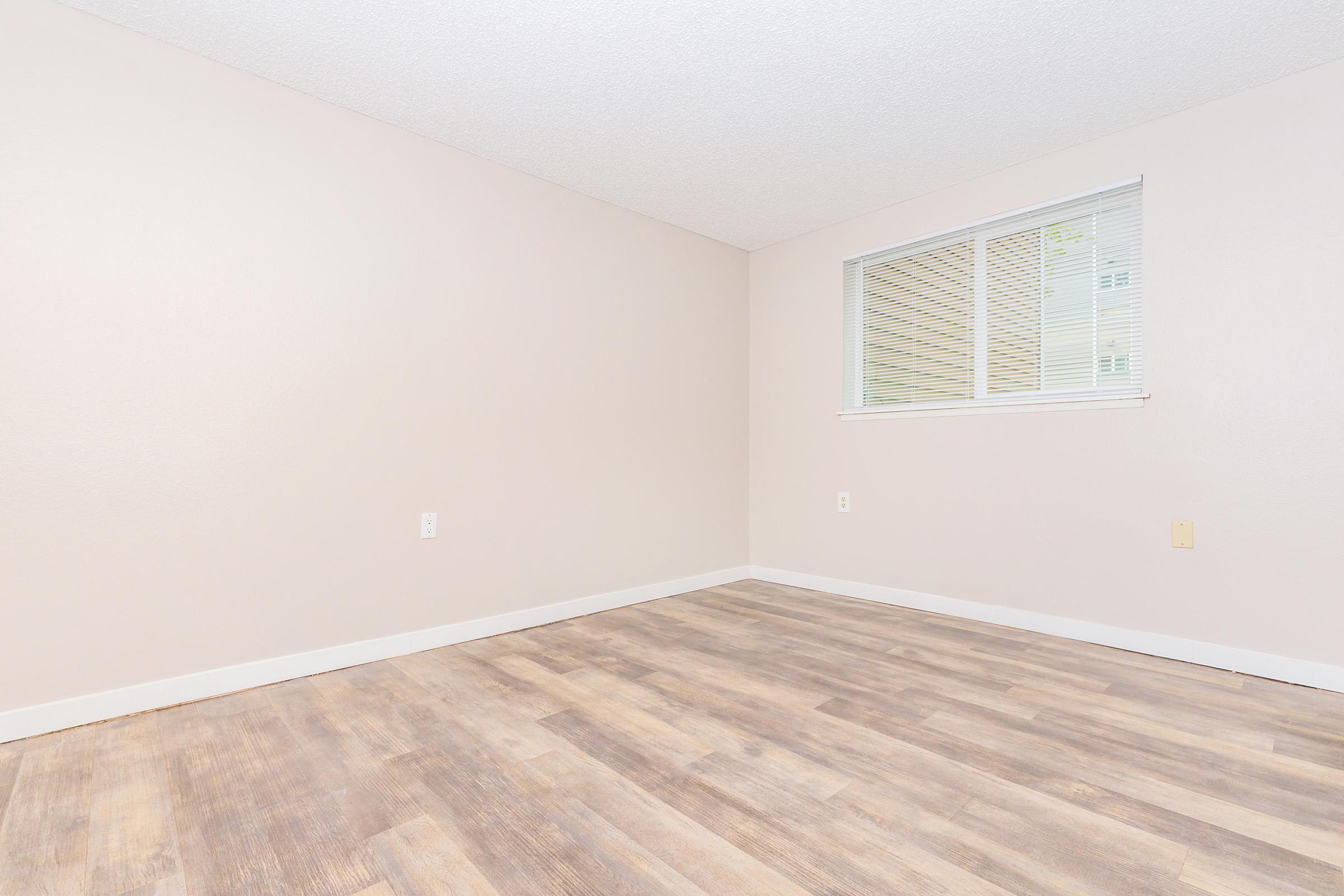 An empty room with light beige walls and a window on one side. The flooring is a light wood laminate. There are no furniture or decorations, creating a spacious and minimalist feel. The lighting is bright and neutral.