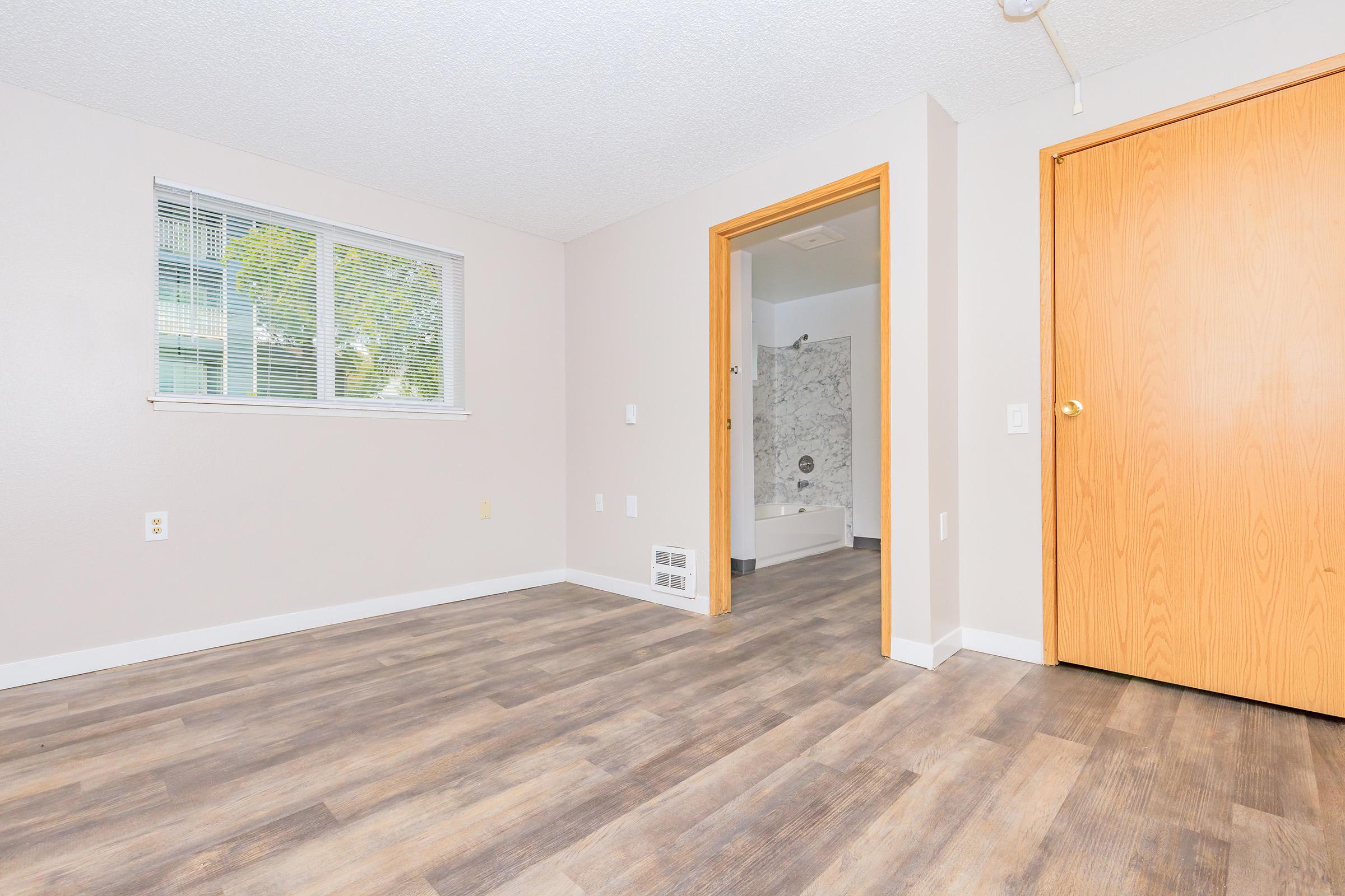 A vacant room with light gray walls and wood laminate flooring. A window on the left lets in natural light. There's an open door on the right leading to a bathroom area with marble walls and a tub visible. The room has a clean, modern appearance, suitable for various uses.