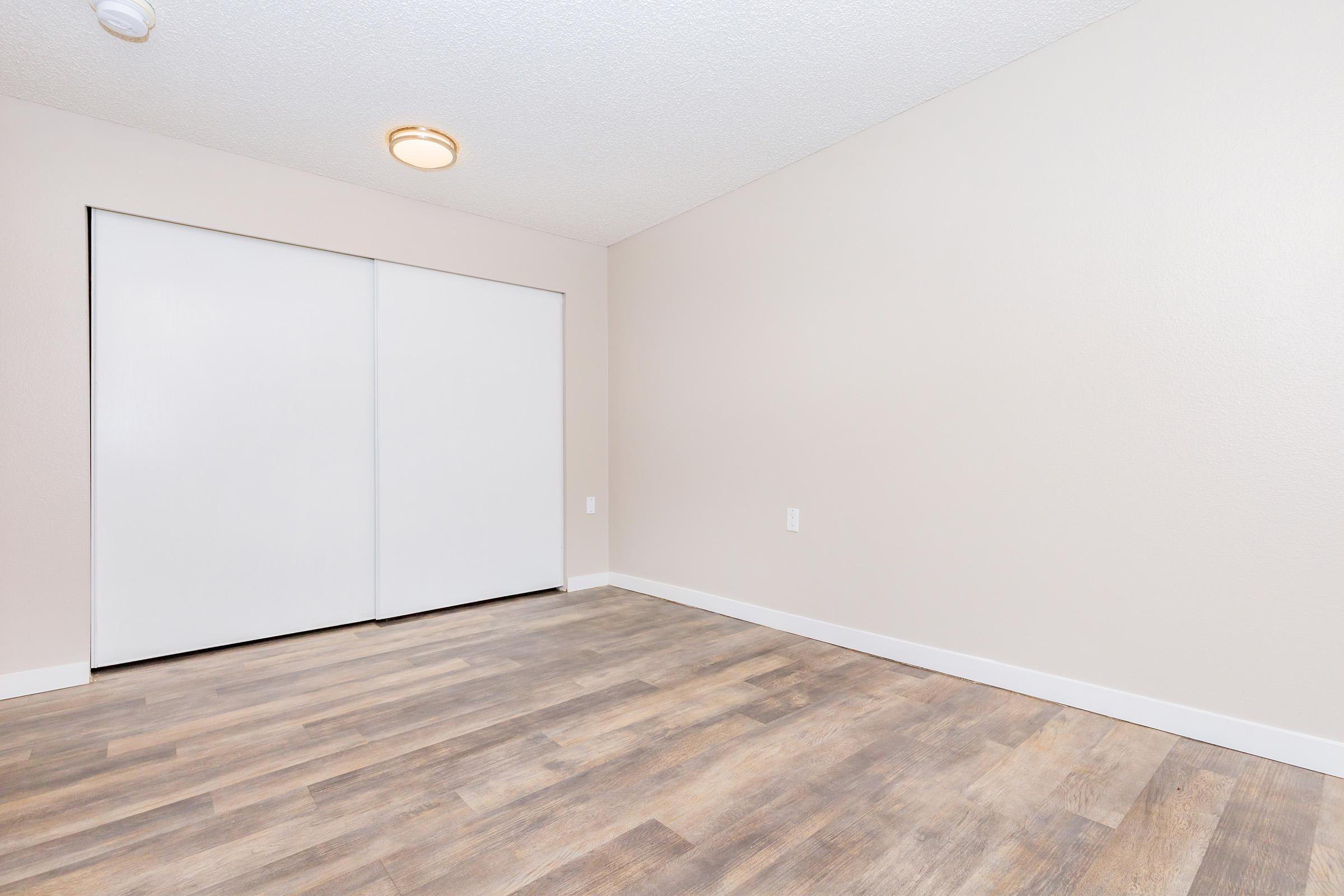 A light-colored room with a textured ceiling, featuring a large closet with white sliding doors. The floor is made of wood-like laminate, and the walls are painted a neutral beige. There is a ceiling light fixture, and the space appears clean and unfurnished.
