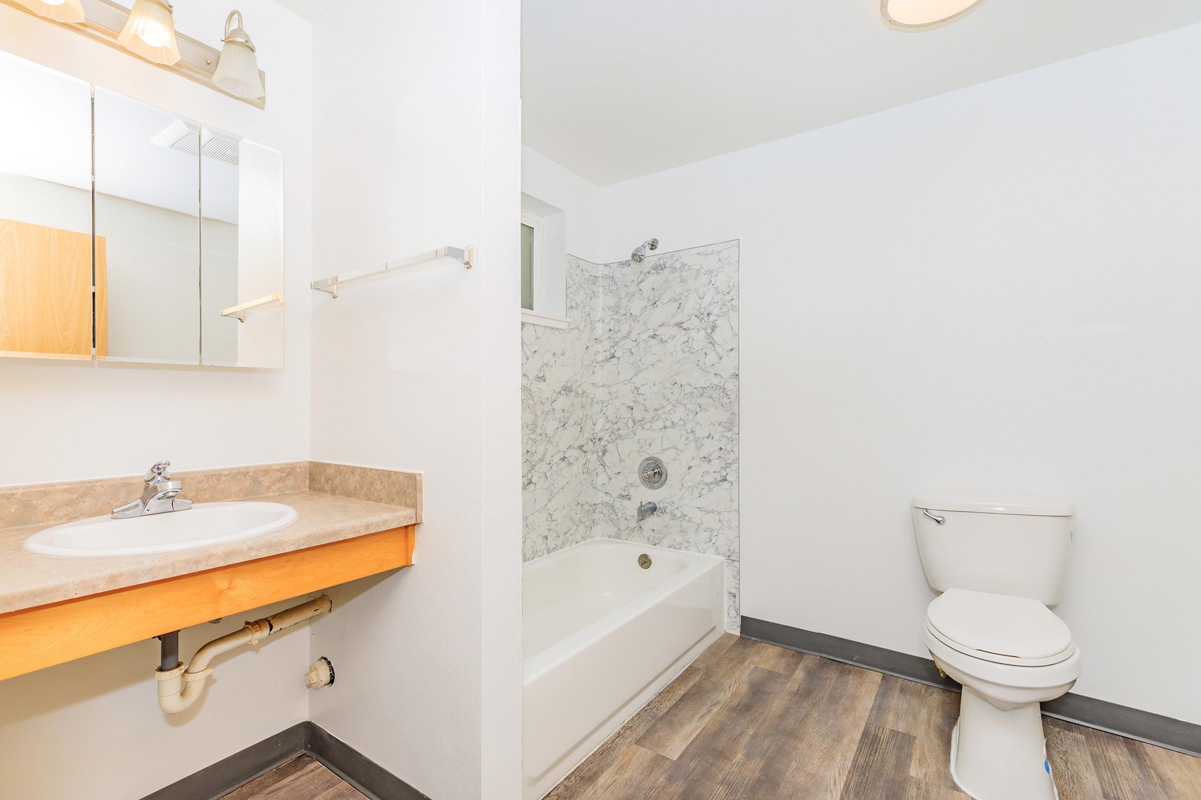 A clean and modern bathroom featuring a white bathtub with a marble surround, a toilet, a sink with a wooden vanity, and a wall-mounted mirror. The floor has a wooden appearance, and the walls are painted white, creating a bright and spacious atmosphere.