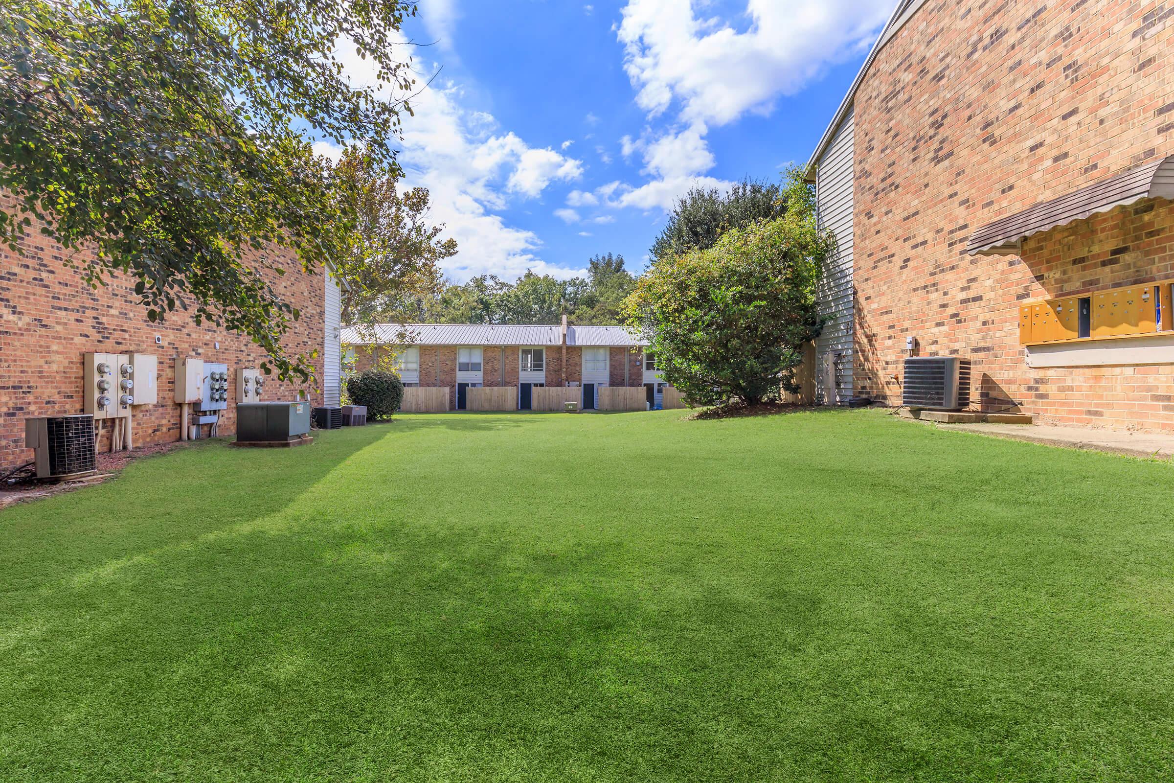A well-maintained green lawn surrounded by brick buildings under a blue sky with scattered clouds. The scene features air conditioning units on the sides of the buildings and trees providing some shade, creating a peaceful outdoor space.