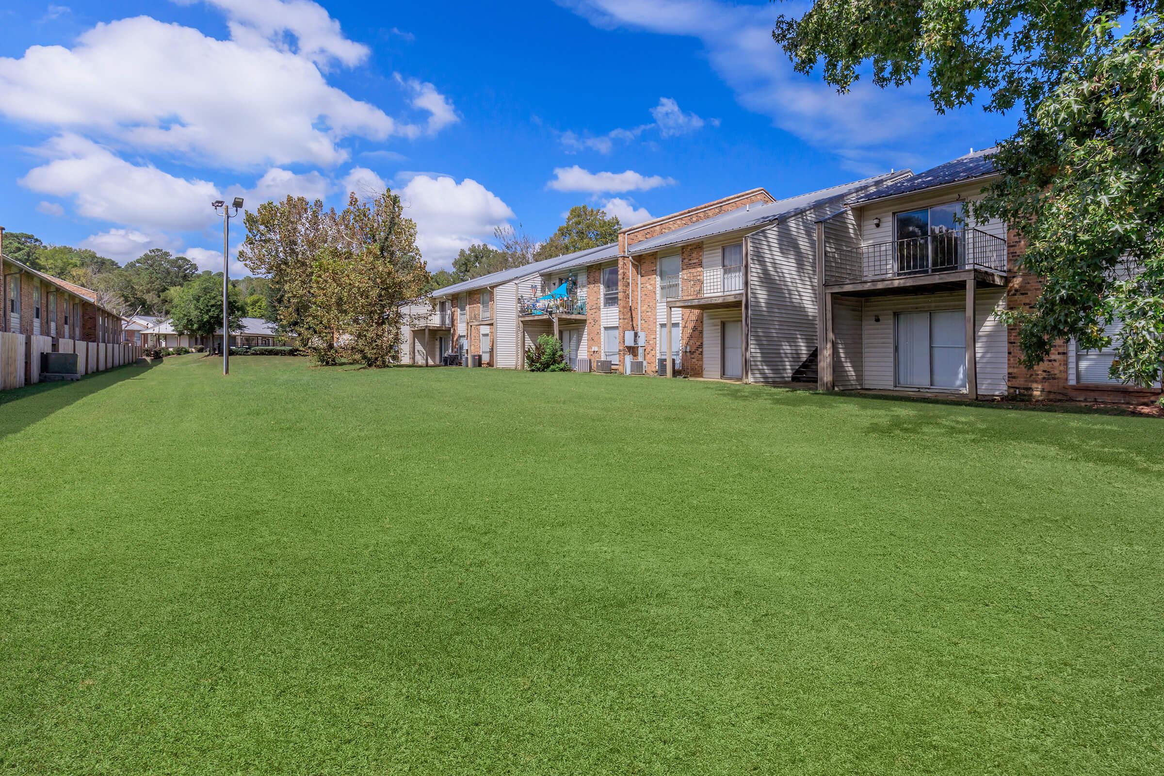 A sunny day overlooks a well-maintained green lawn with several multi-level apartment buildings, featuring brick and wooden exteriors. The sky is partly cloudy, adding to the inviting atmosphere of the residential area.
