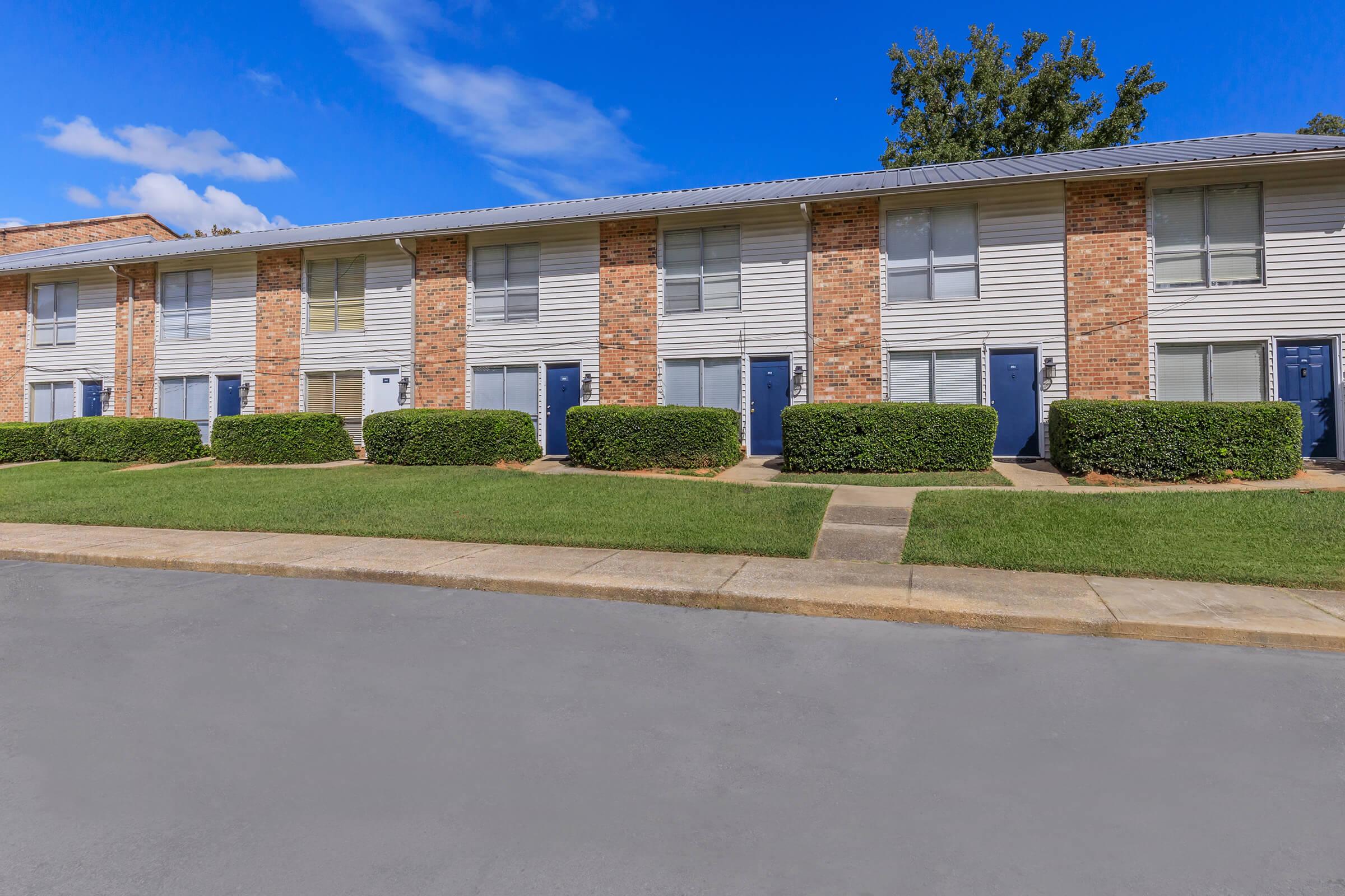 A row of two-story apartment buildings with brick and siding exteriors. Each unit features blue doors and manicured hedges in front. The sky is clear with a few clouds, and the parking area is visible in the foreground, surrounded by well-kept grass.