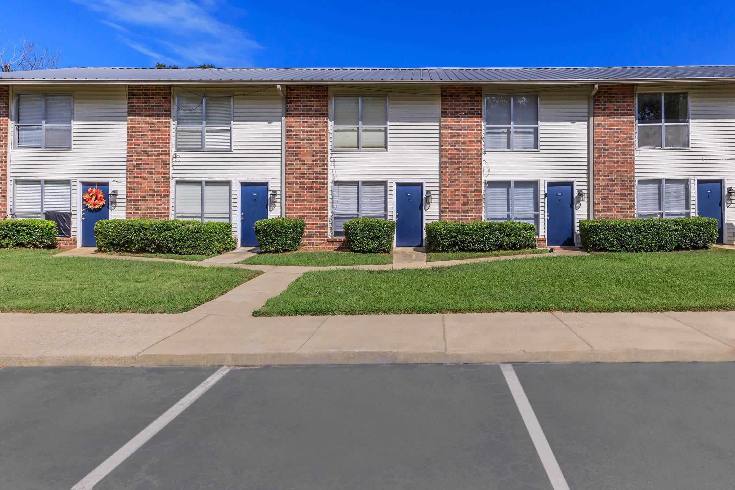 Exterior view of a multi-unit residential building with brick and siding walls. Each unit has a front door labeled with a number. Neatly trimmed bushes line the walkway, and green grass is visible in the foreground. The sky is clear and blue, indicating a sunny day.