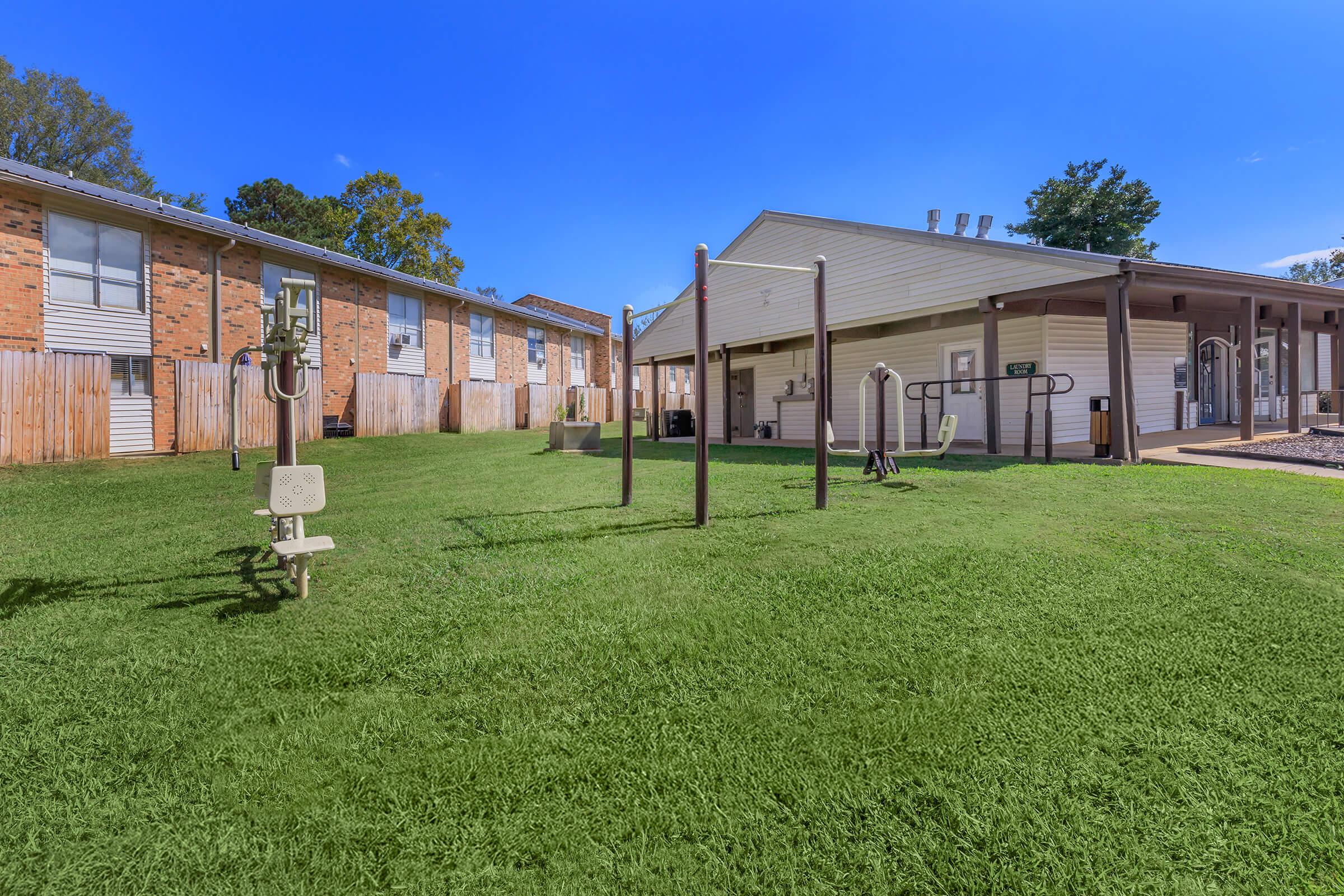 Outdoor fitness area featuring exercise equipment on green grass, with two buildings in the background. Clear blue sky above adds to the bright atmosphere. The space is well-maintained, inviting for physical activity and community engagement.