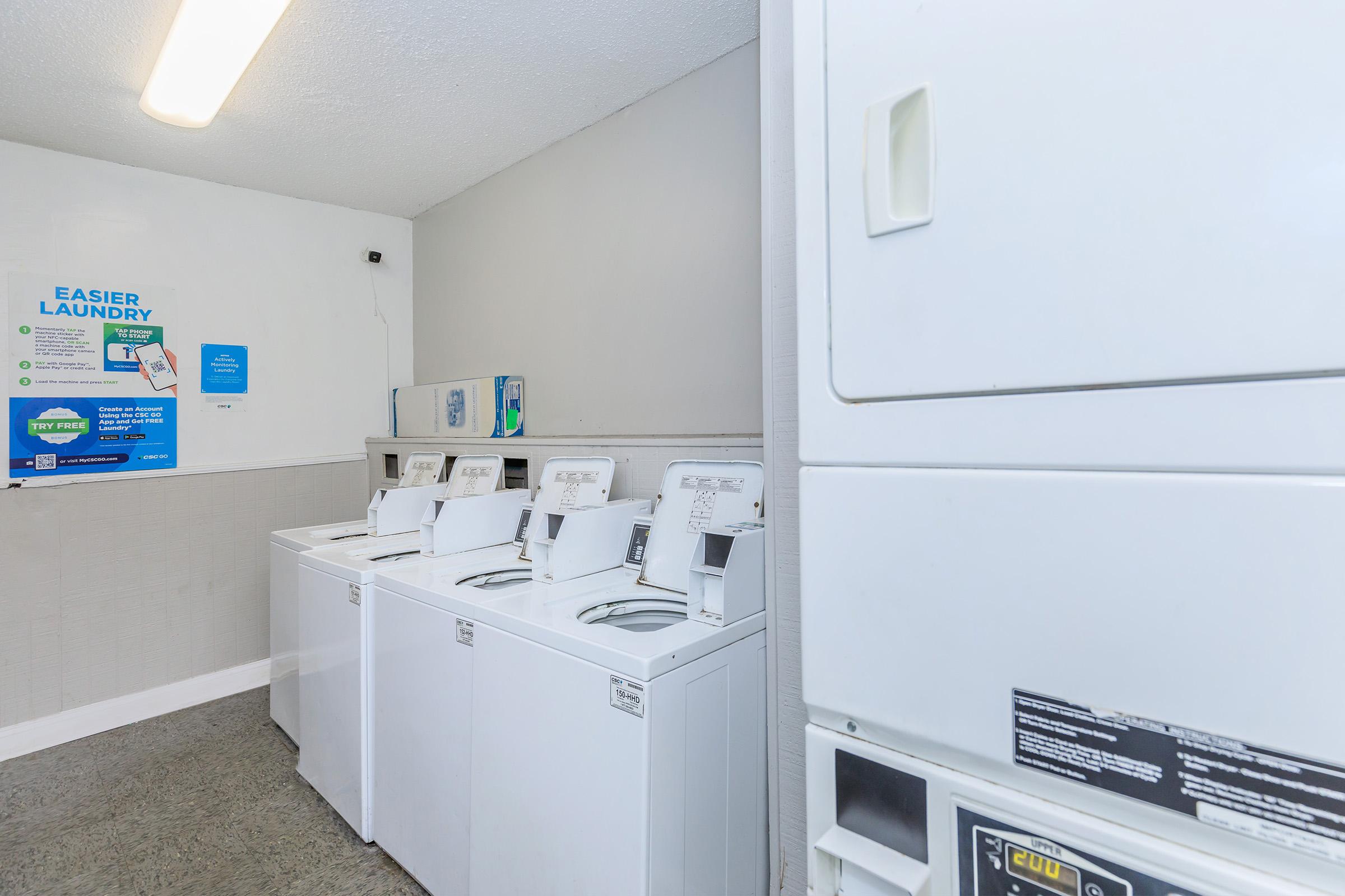 A clean and well-lit laundry room featuring several white washing machines and a dryer. The wall is painted light gray, and a poster with laundry instructions is visible on the left. The floor is a neutral color, complementing the overall tidy appearance of the space.