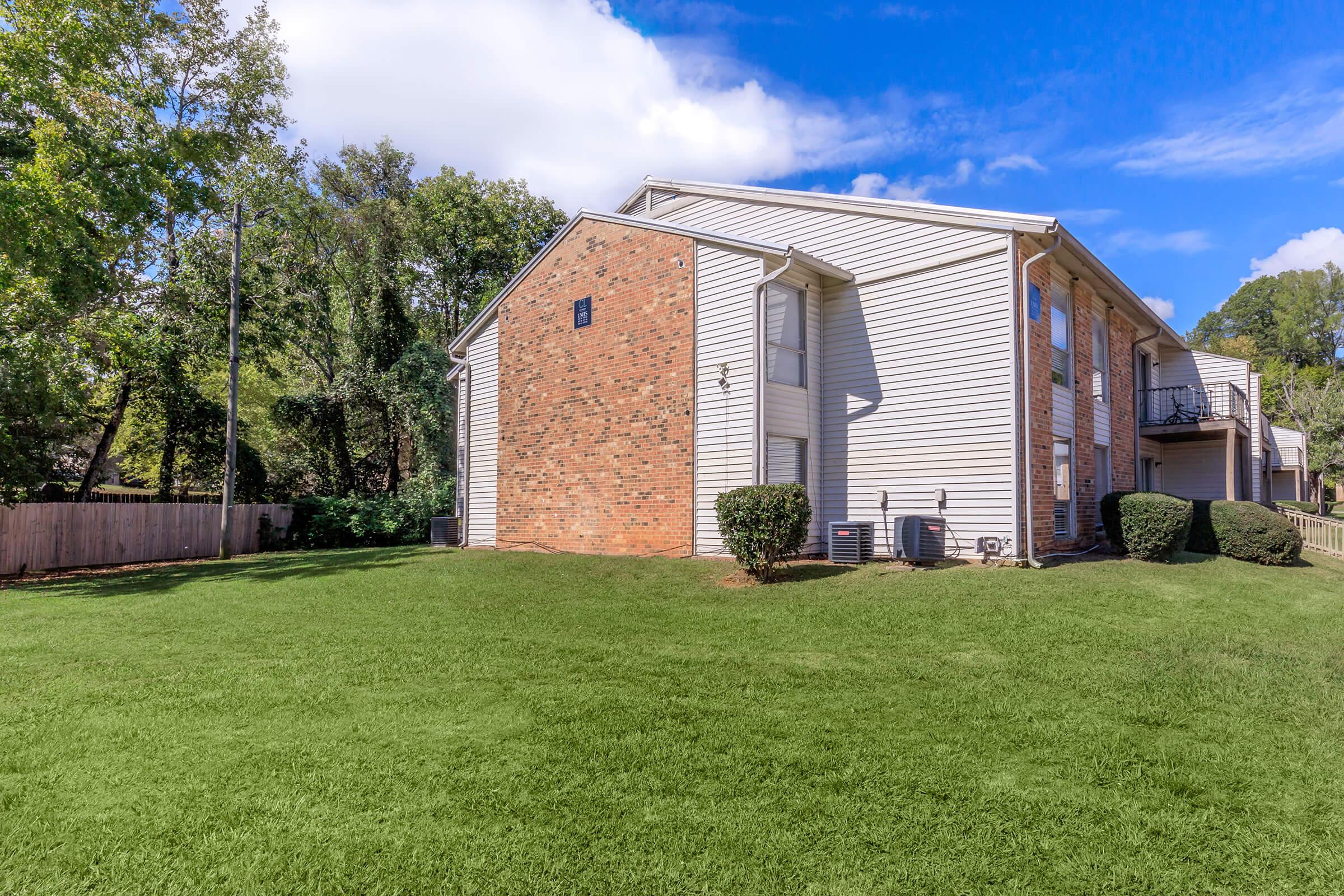 The image shows the side of a two-story apartment building, featuring a mix of brick and siding. It is surrounded by a well-maintained lawn with neatly trimmed bushes and a clear blue sky with a few clouds in the background.