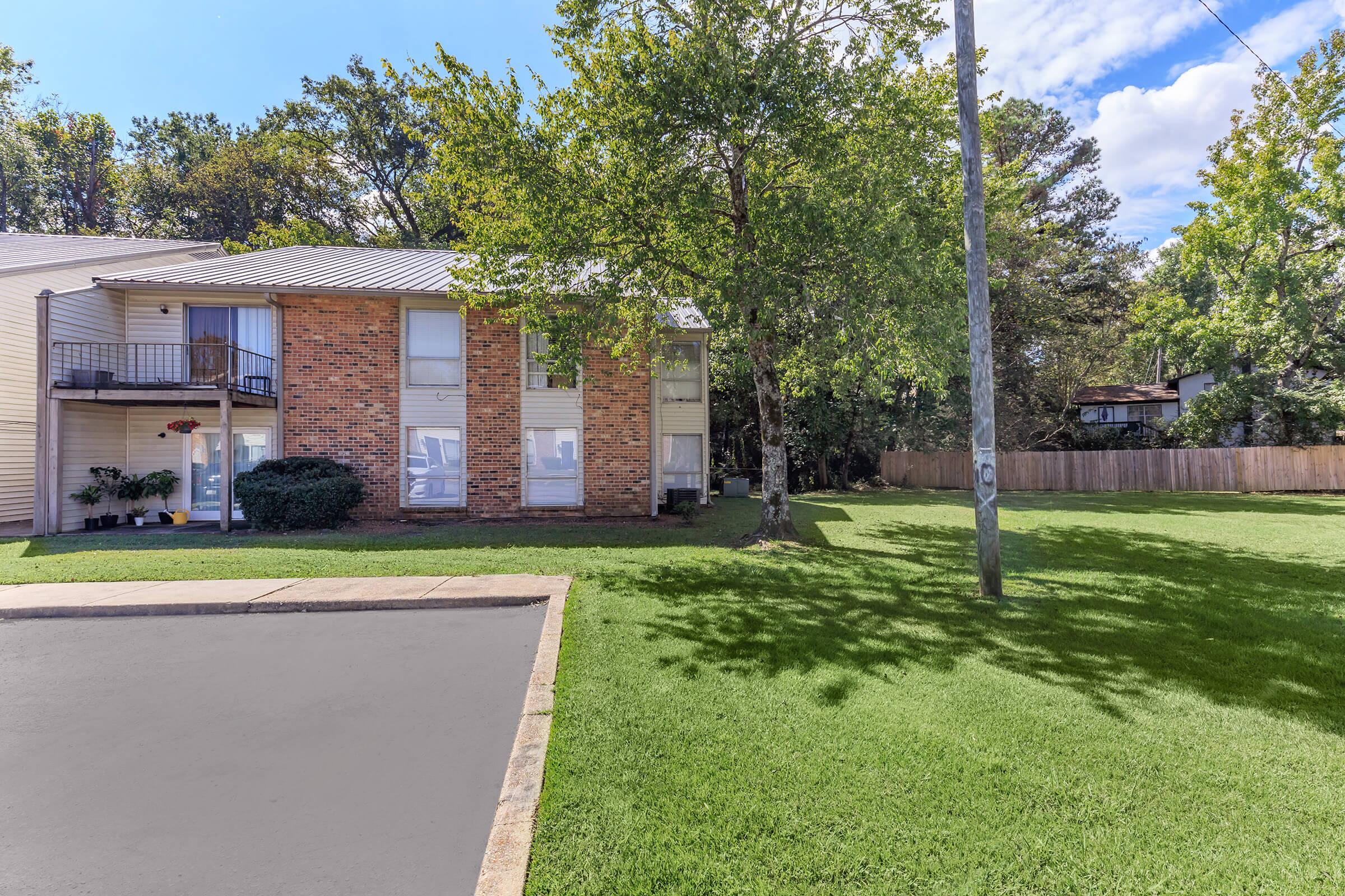 Exterior view of a two-story brick apartment building surrounded by green grass and trees. A power pole stands nearby, and there is a parking lot in front. The sky is partly cloudy, creating a bright atmosphere. A fenced backyard is visible in the background.