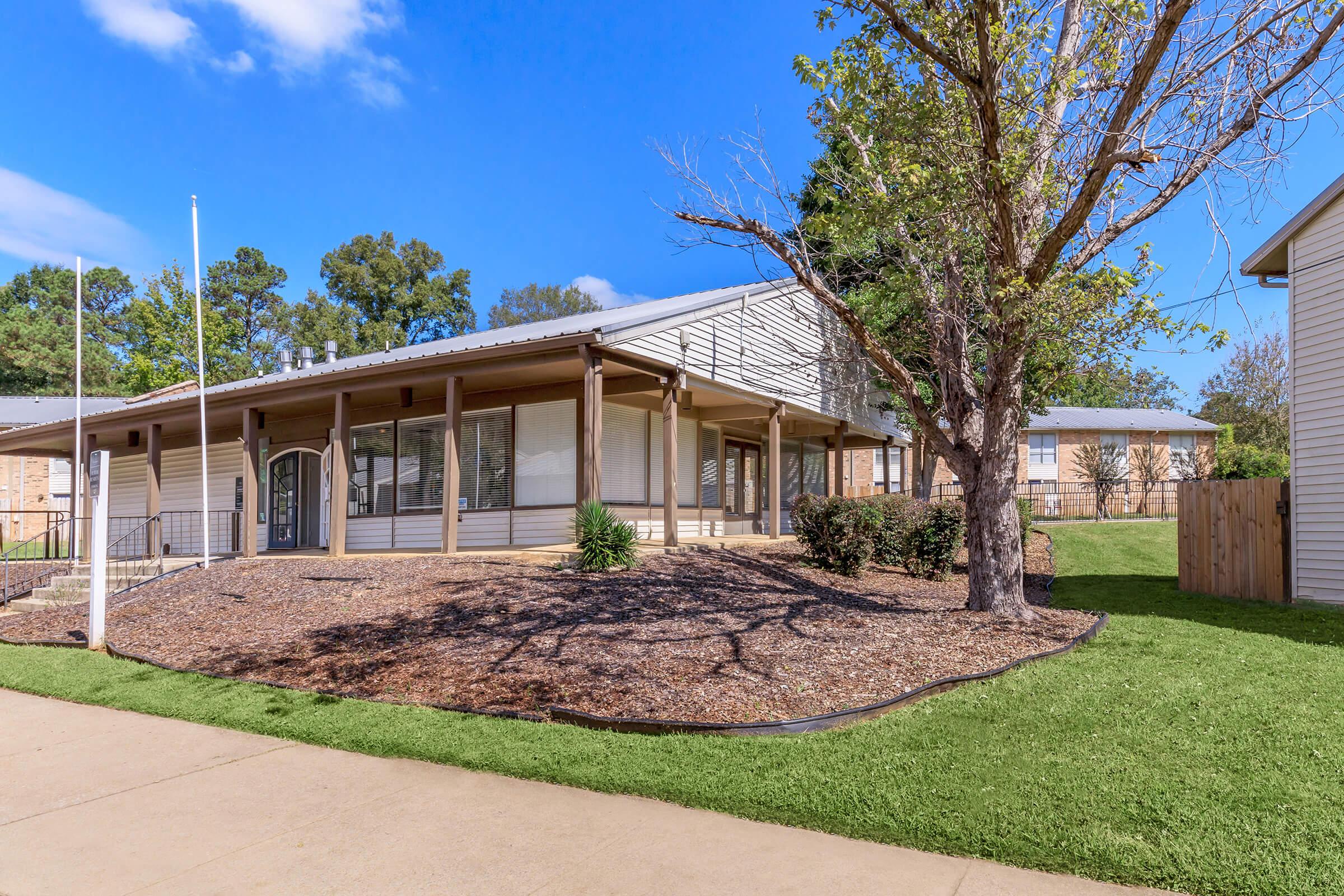 A modern, single-story building surrounded by landscaped grounds, including a grassy area and a tree. The facade features a wide porch, large windows, and a mix of siding materials. Blue sky and a few clouds enhance the bright, inviting atmosphere.